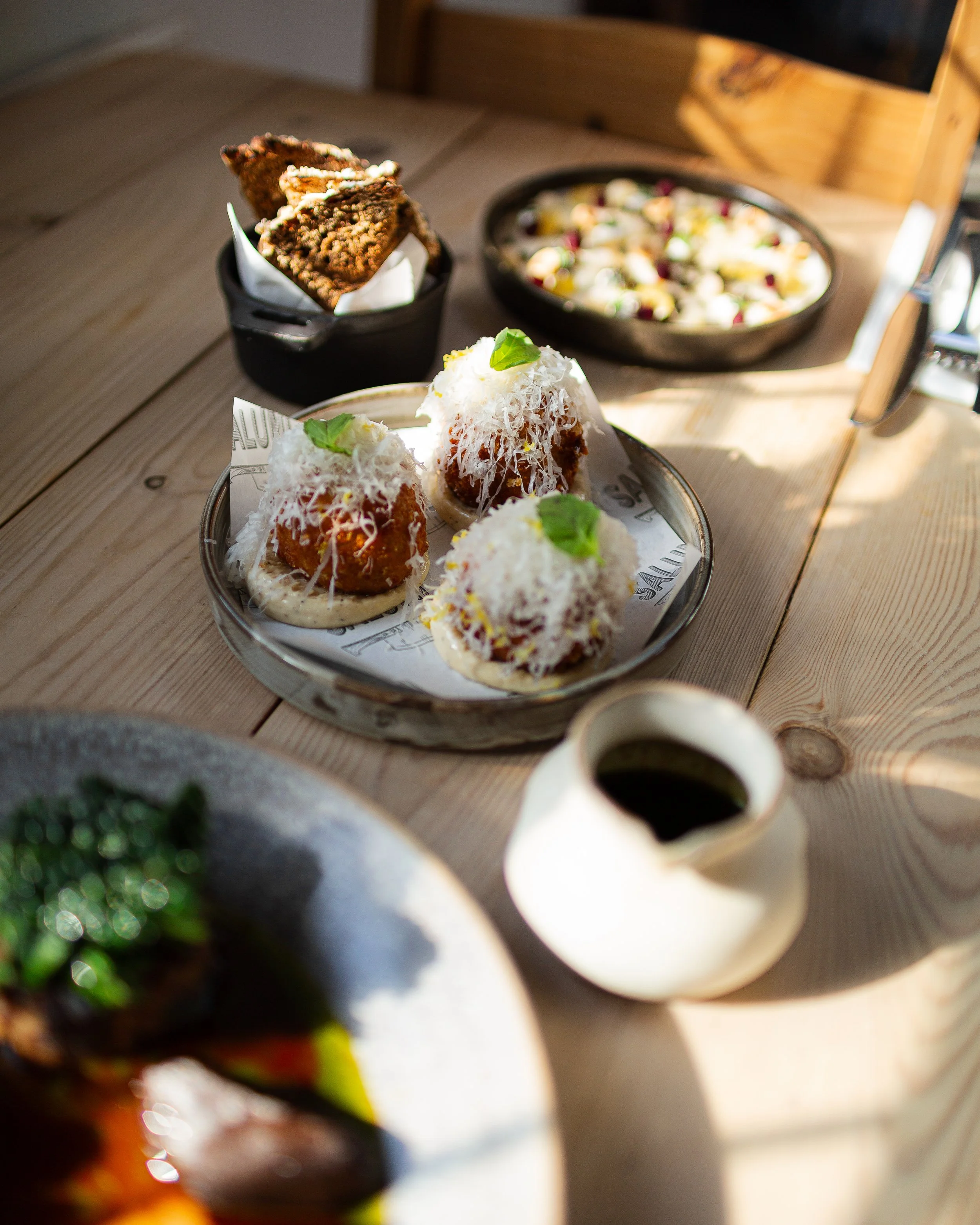 Plate of three arancini with grated cheese and basil on top, a bowl of fried cutlets or croquettes, a bowl of salad with cheese and vegetables, and a small pitcher of dipping sauce on a wooden table.