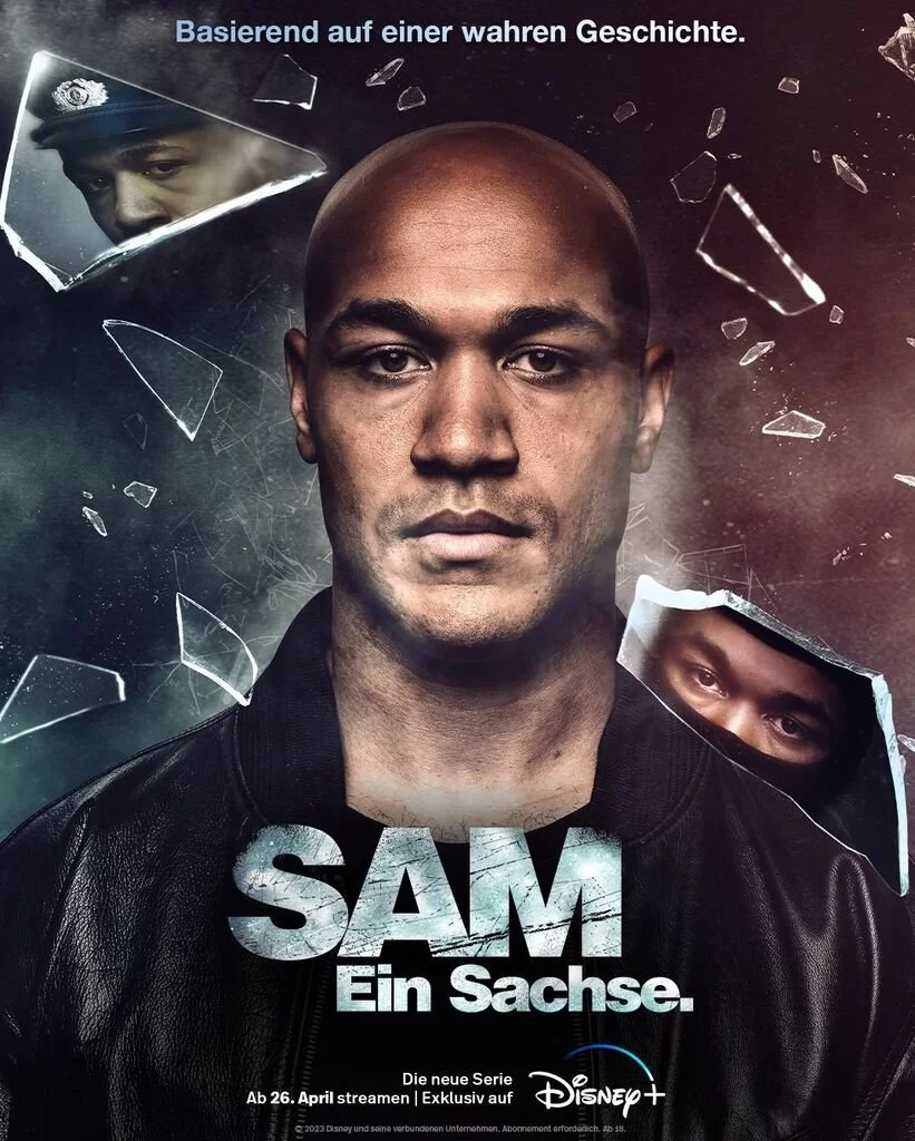 Werbeplakat für eine neue Serie namens 'SAM - Ein Sachse.' mit einem Hauptbild eines ernst blickenden Mannes in Lederjacke, umgeben von zerbrochenem Glas und zwei kleine Gesichter, die durch Löcher im Glas sichtbar sind, einem auf der linken Seite, d