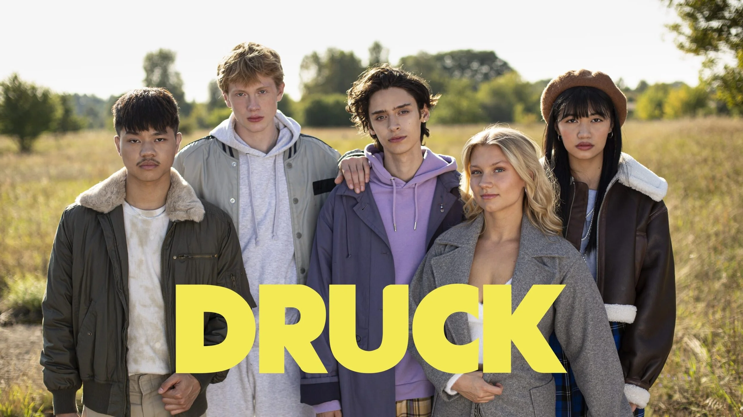 Eine Gruppe von sechs jungen Menschen steht im Freien, Sonnenlicht, im Hintergrund Bäume und ein Feld, mit gelbem Text 'DRUCK' auf dem Bild.