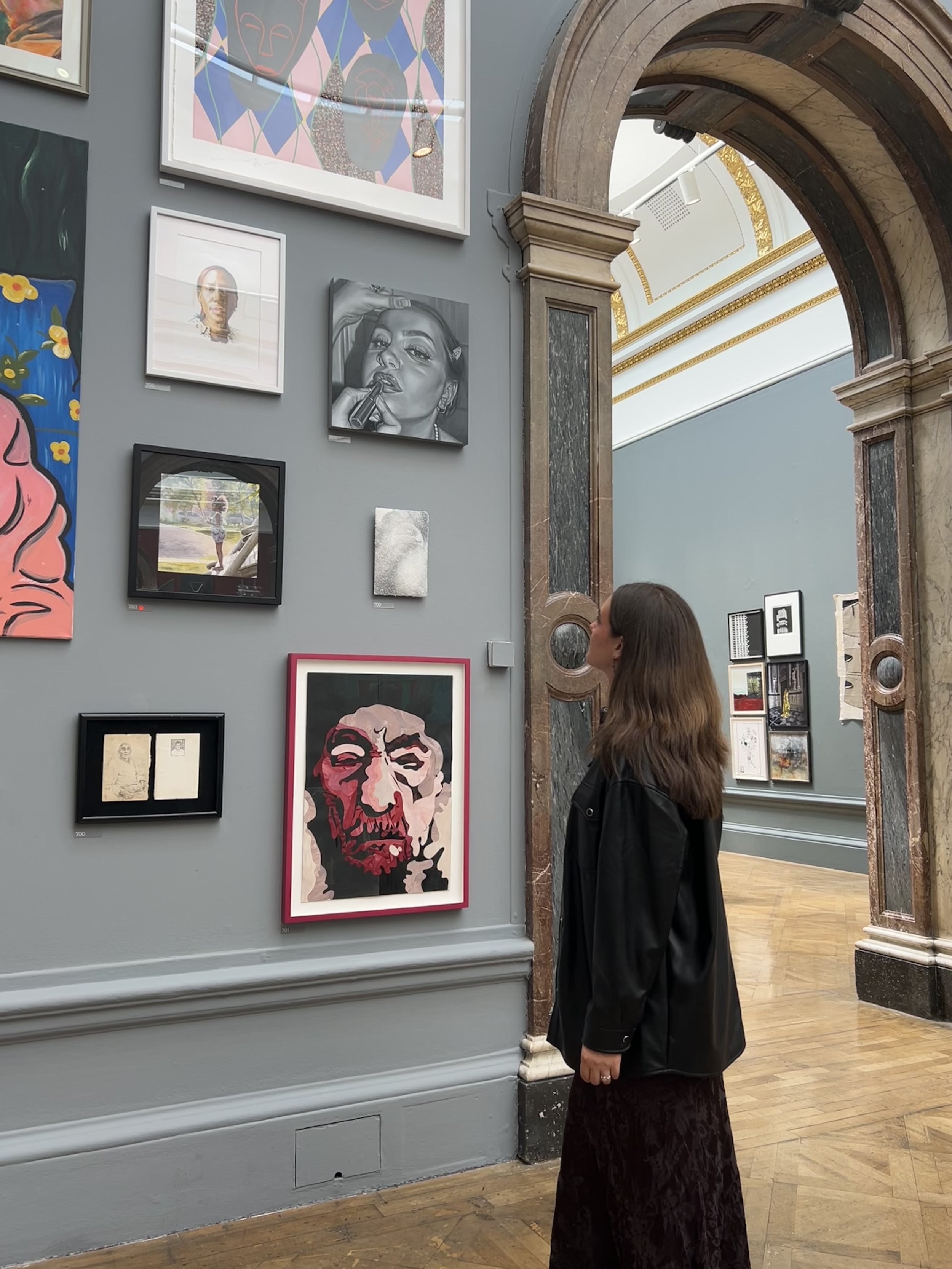 A woman with long brown hair, wearing a black jacket and a dark long skirt, stands in an art gallery looking at various artworks on a gray wall, including portraits, a pencil sketch, and a colorful abstract piece.