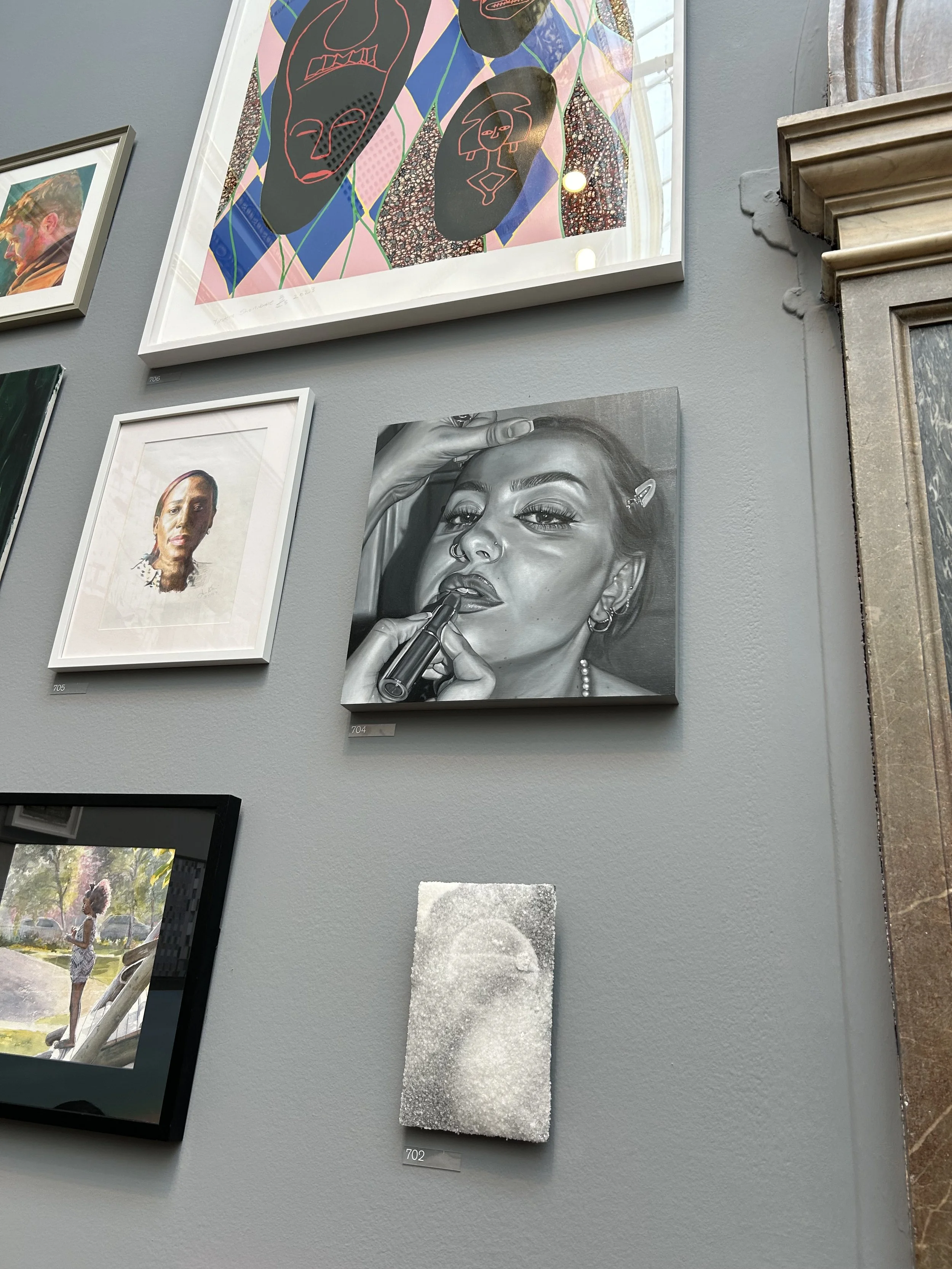 A gallery wall displaying various artworks, including colorful abstract illustrations, a realistic grayscale portrait of a woman with makeup and jewelry, a digital artwork of a person standing outdoors, and a mixed media piece resembling a textured face. The artworks are framed and mounted on a gray wall.