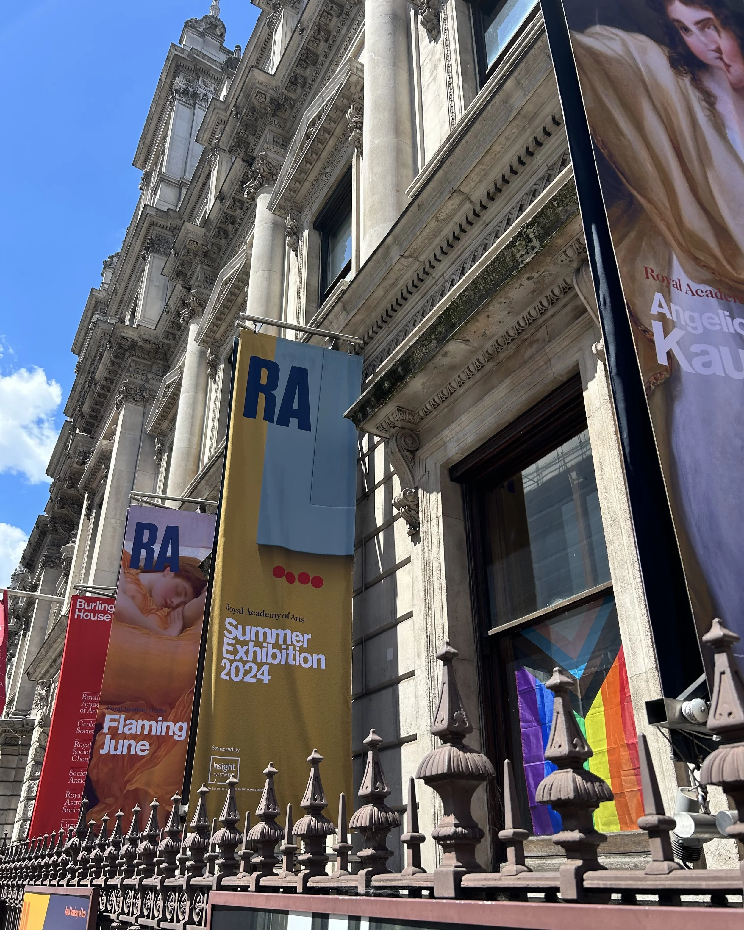 Exterior view of a historic building with ornate architecture, displaying colorful banners for the Royal Academy of Arts Summer Exhibition 2024, including one with a rainbow flag, and a partial rainbow pride flag inside the building next to a window.