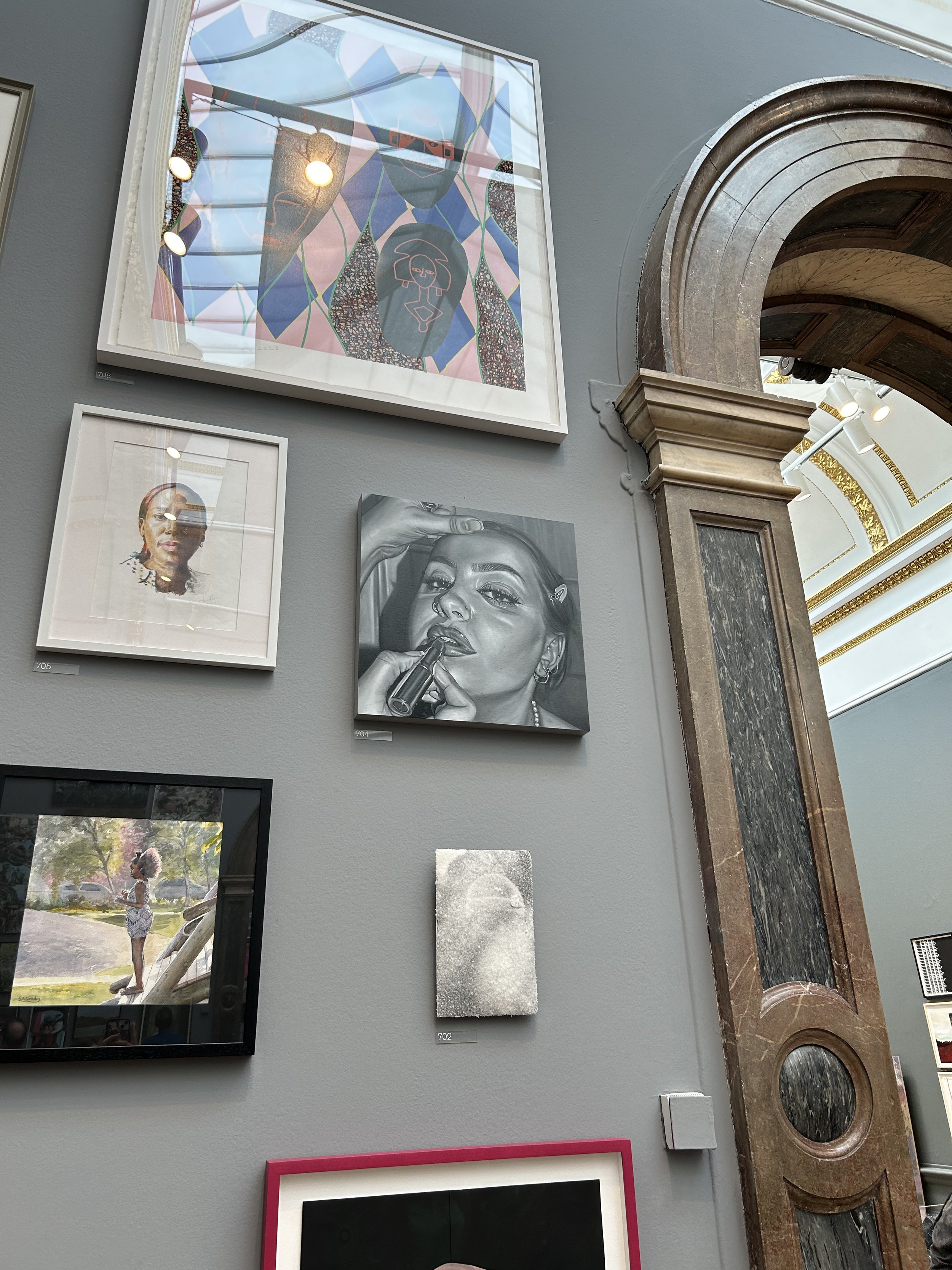 Assorted framed artworks on a gray gallery wall, including a colorful abstract portrait, a realistic black-and-white portrait of a woman with a lipstick, and a photograph of a girl standing outdoors.