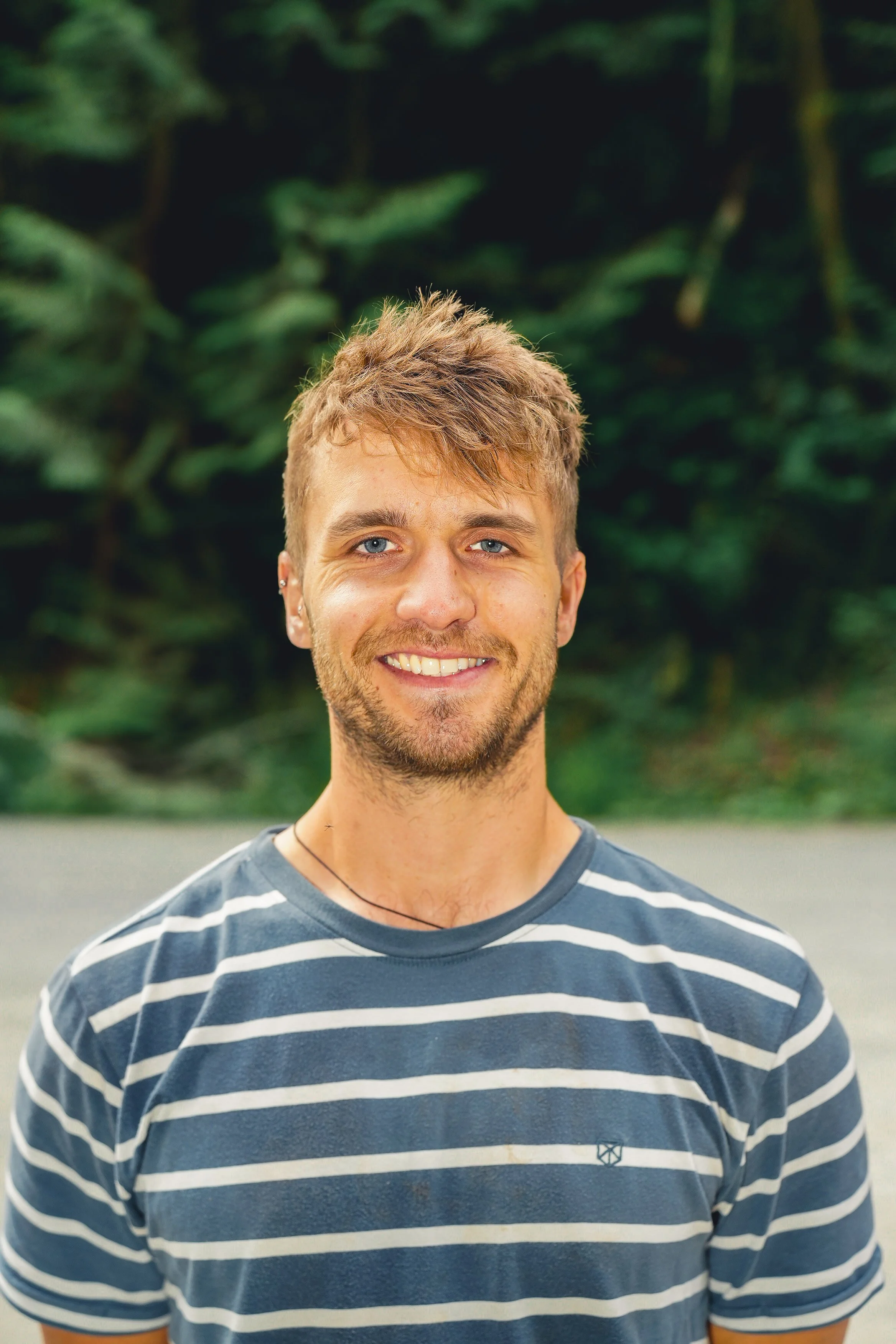 Joe Wakefield headshot. light brown hair and a beard, smiling, wearing a blue and white striped t-shirt, standing outdoors with green trees in the background.