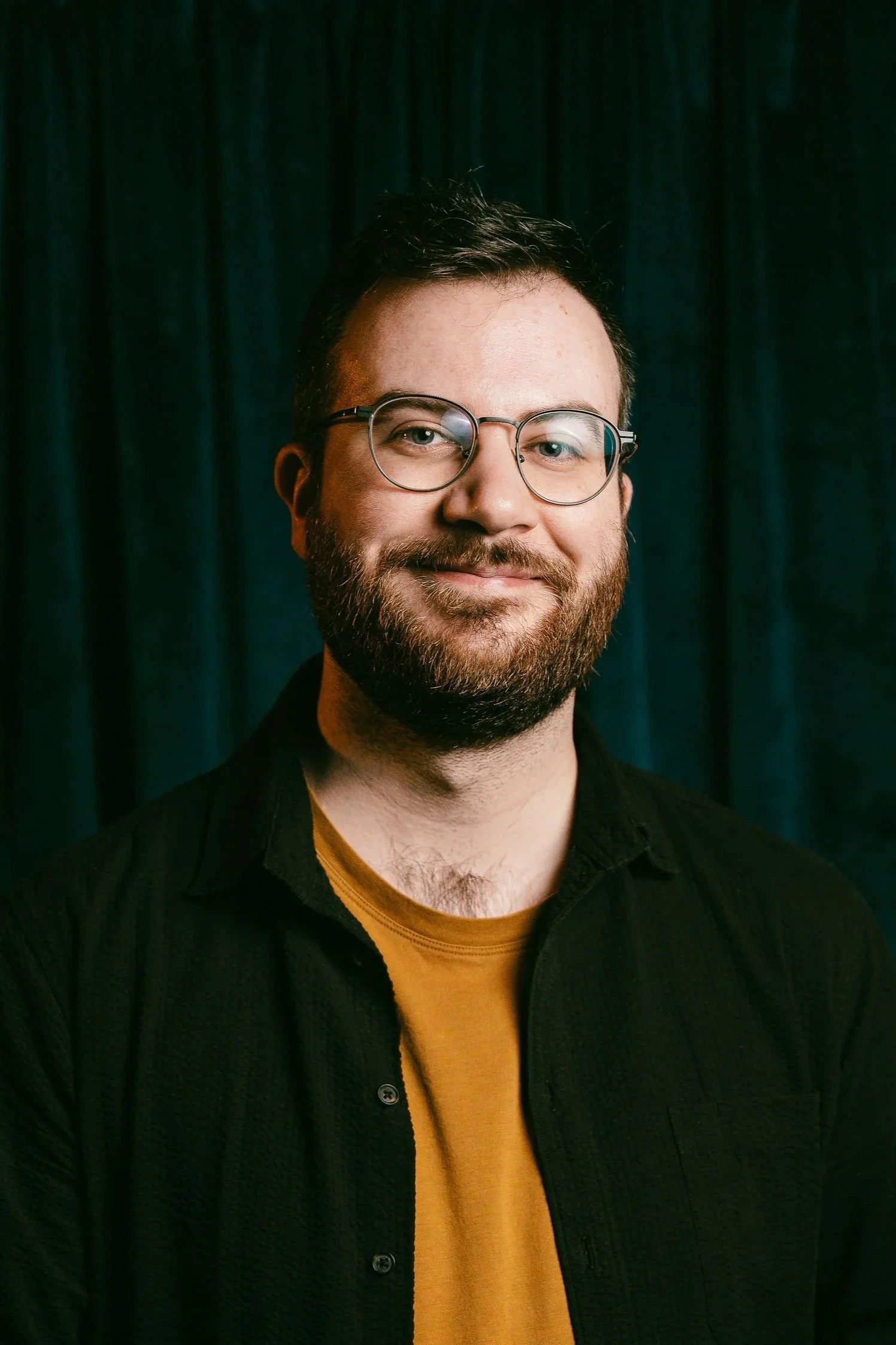 Portrait of a smiling man with glasses, a beard, and short dark hair, wearing a black shirt and a mustard-colored T-shirt, against a dark green curtain background.