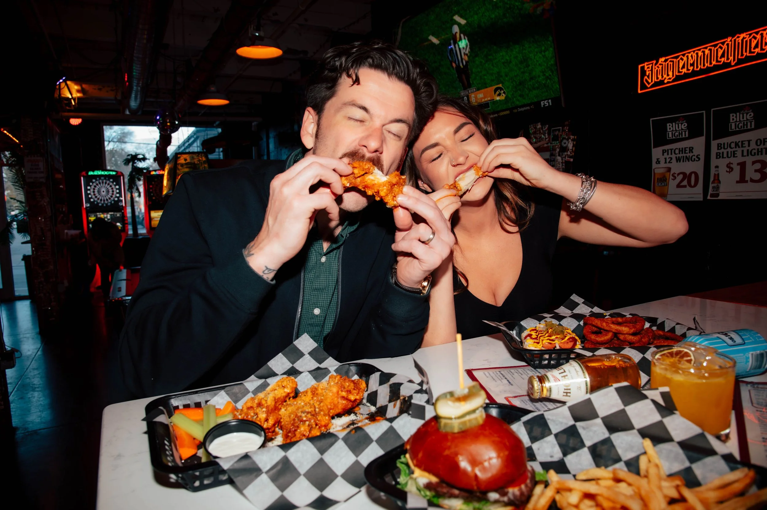 Two people enjoying chicken wings and fries at a restaurant with arcade games in the background.