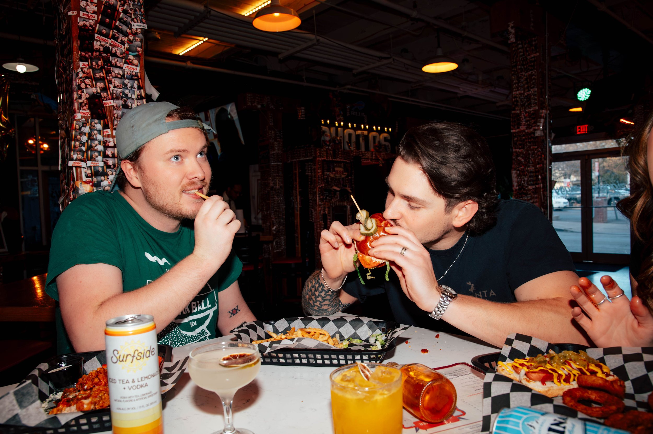 Two men eating at a restaurant table with drinks and food, one is holding a burger and the other is eating a fry, with a busy restaurant interior in the background.