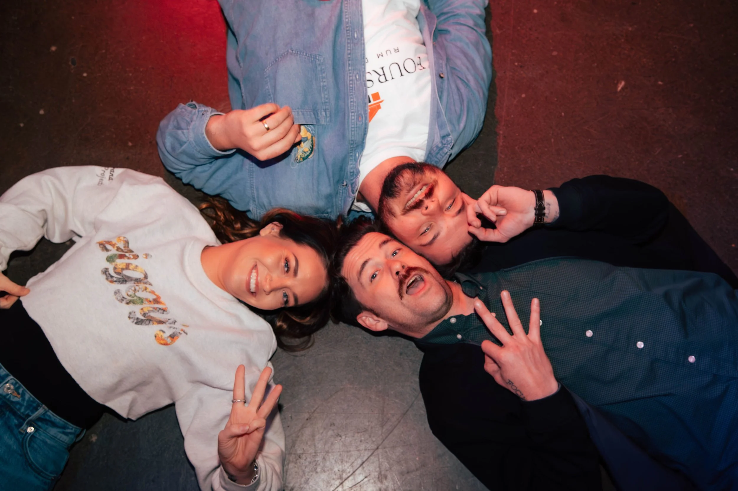 Friends lying on the floor at Ziggy’s neighborhood bar in Rochester, NY, laughing and flashing peace signs during a fun night out wearing Ziggy's merchandise.