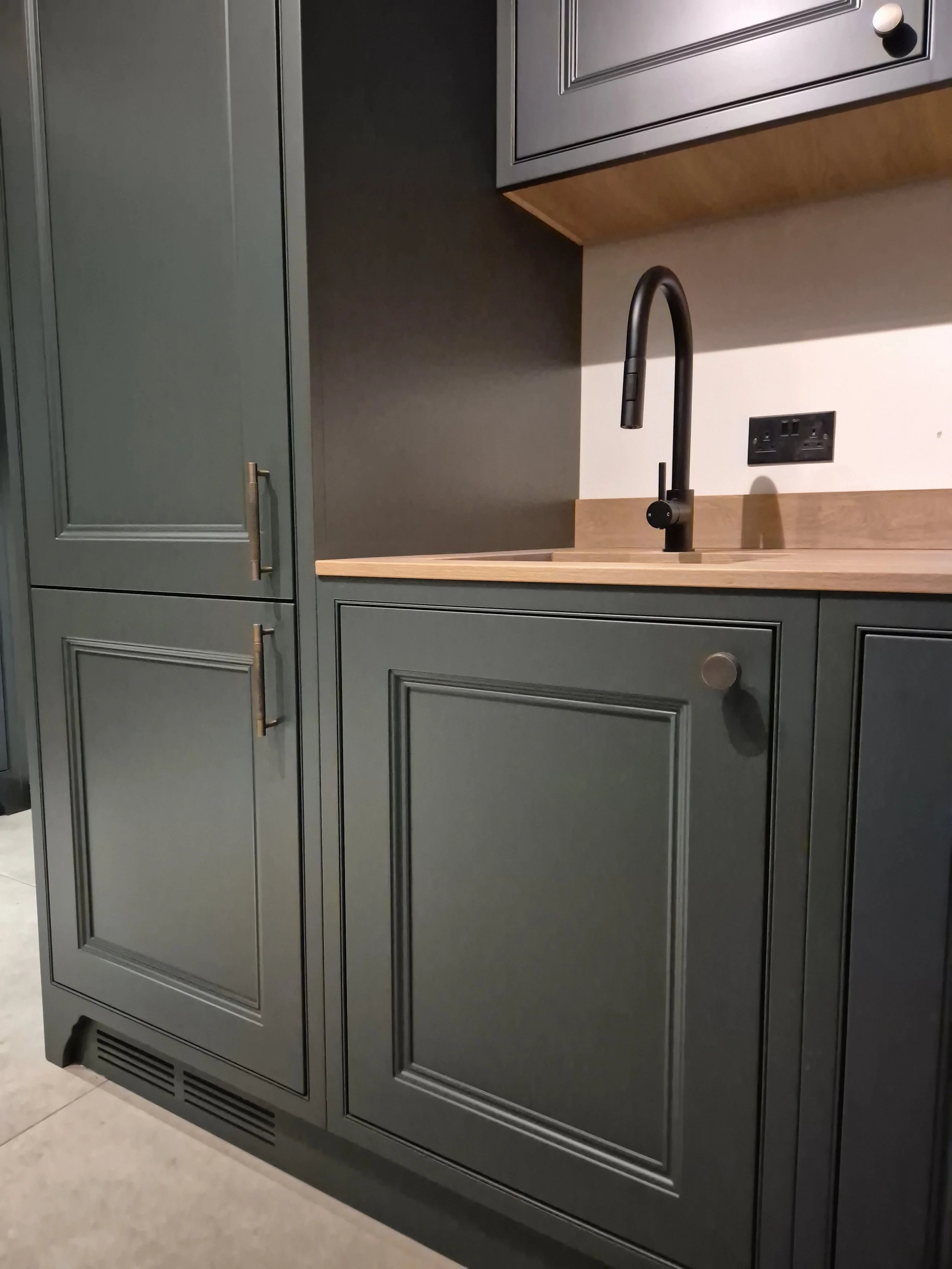 Modern kitchen with dark green cabinetry, light wood countertop, black faucet, and electrical outlets on the wall.