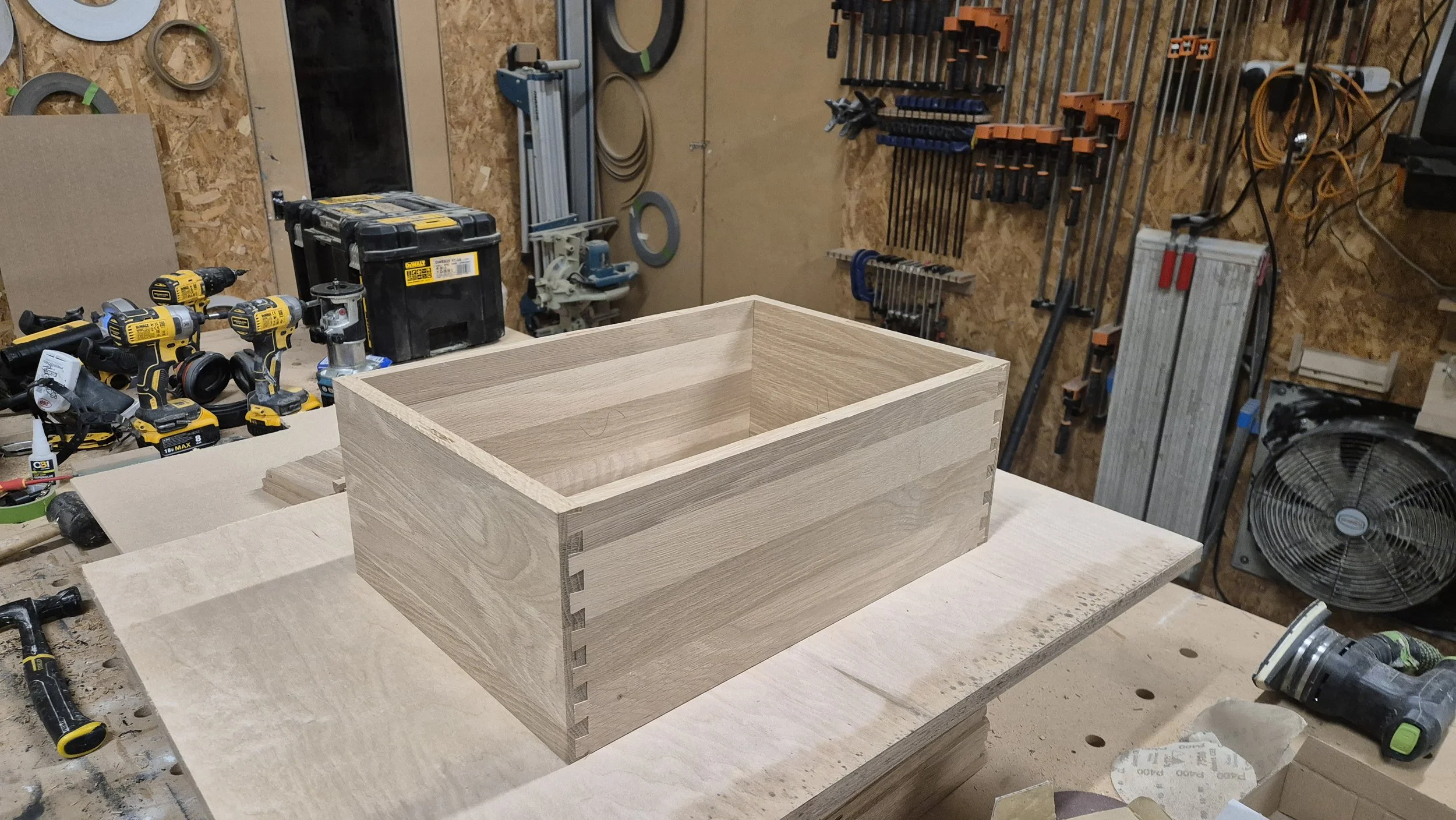 Unfinished wooden box with dovetail joints in a woodworking workshop, surrounded by power tools and clamps.