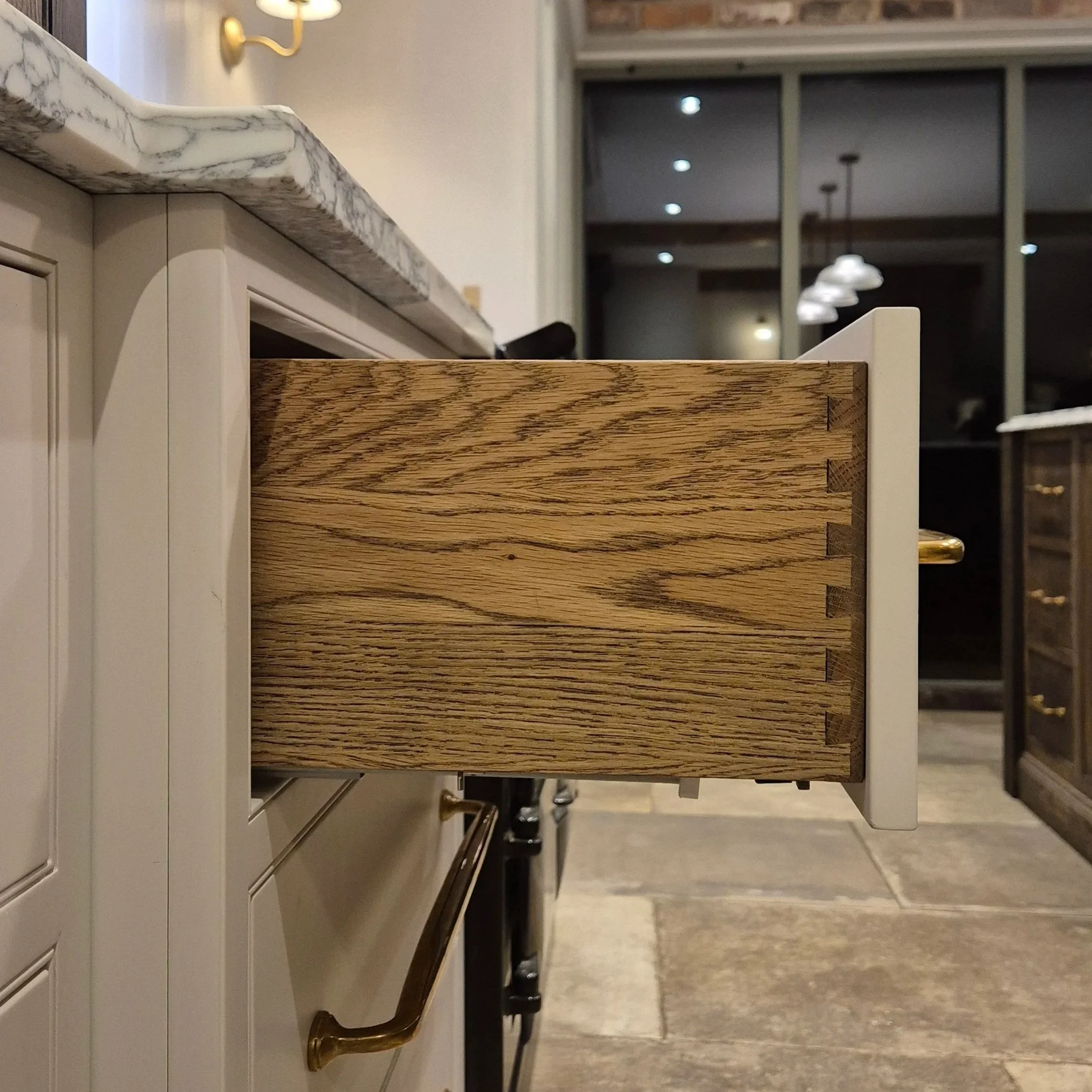Open kitchen drawer with wooden interior, white exterior, in a kitchen with beige cabinets, marble countertop, and dark flooring.