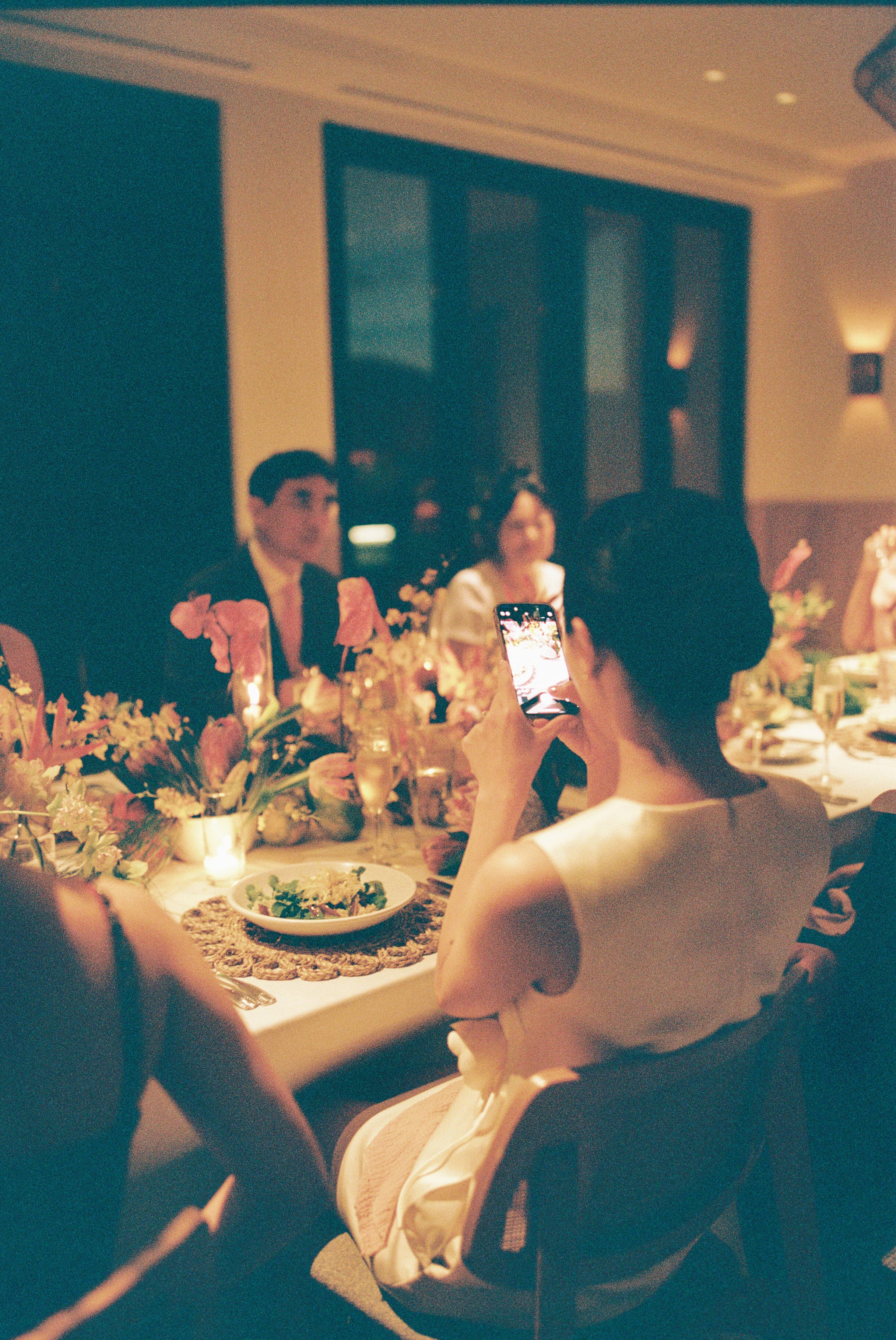 People sitting at a decorated dinner table, one woman taking a photo with her phone, floral arrangements and food on the table, warm ambient lighting.