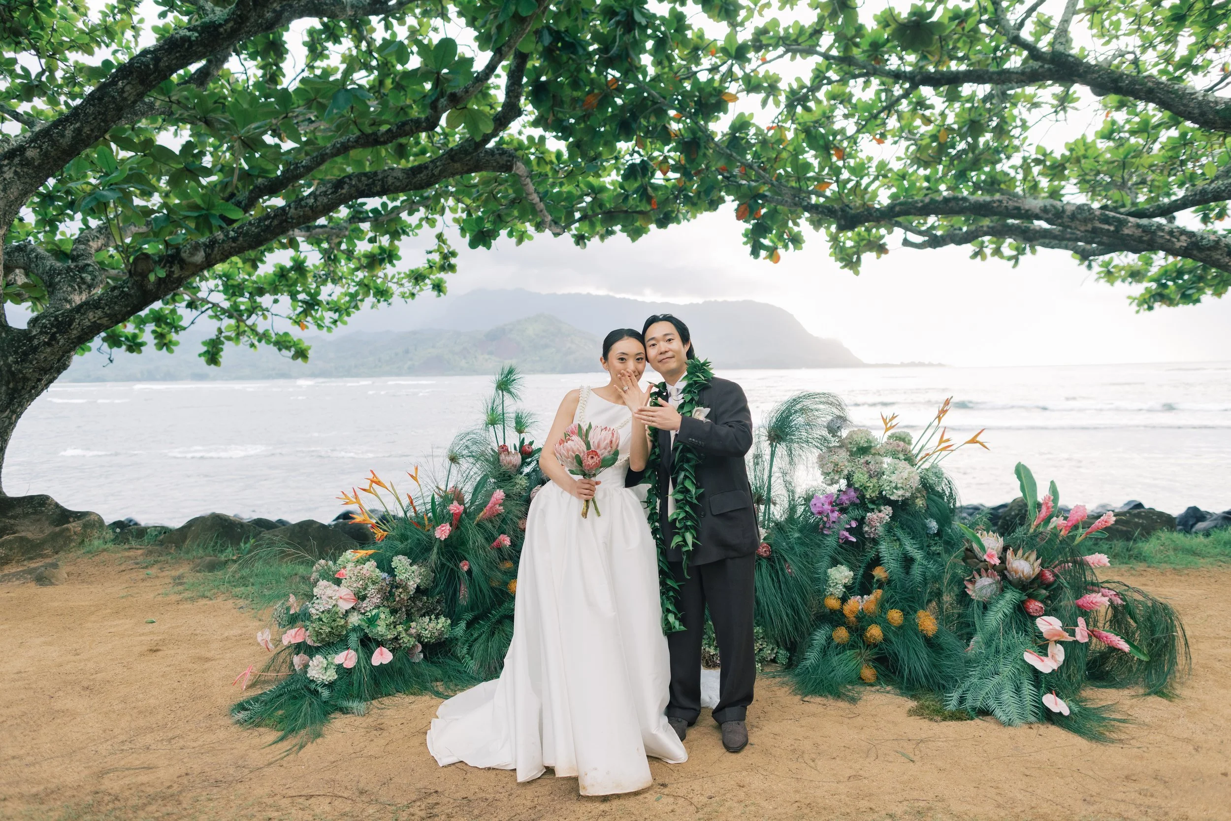 A couple in wedding attire standing on a beach in front of a colorful floral arrangement with a scenic view of water, mountains, and cloudy sky in the background, under a large leafy tree.
