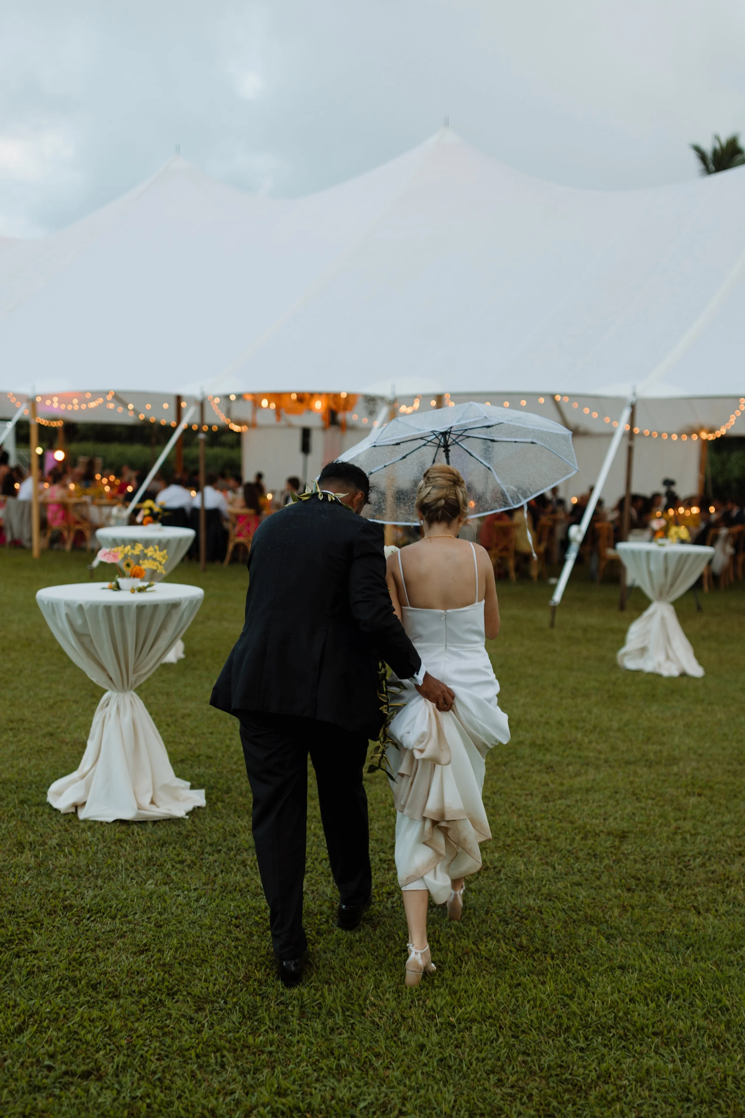 A bride and groom walking under a clear umbrella at an outdoor wedding reception with tables, string lights, and a large white tent in the background.