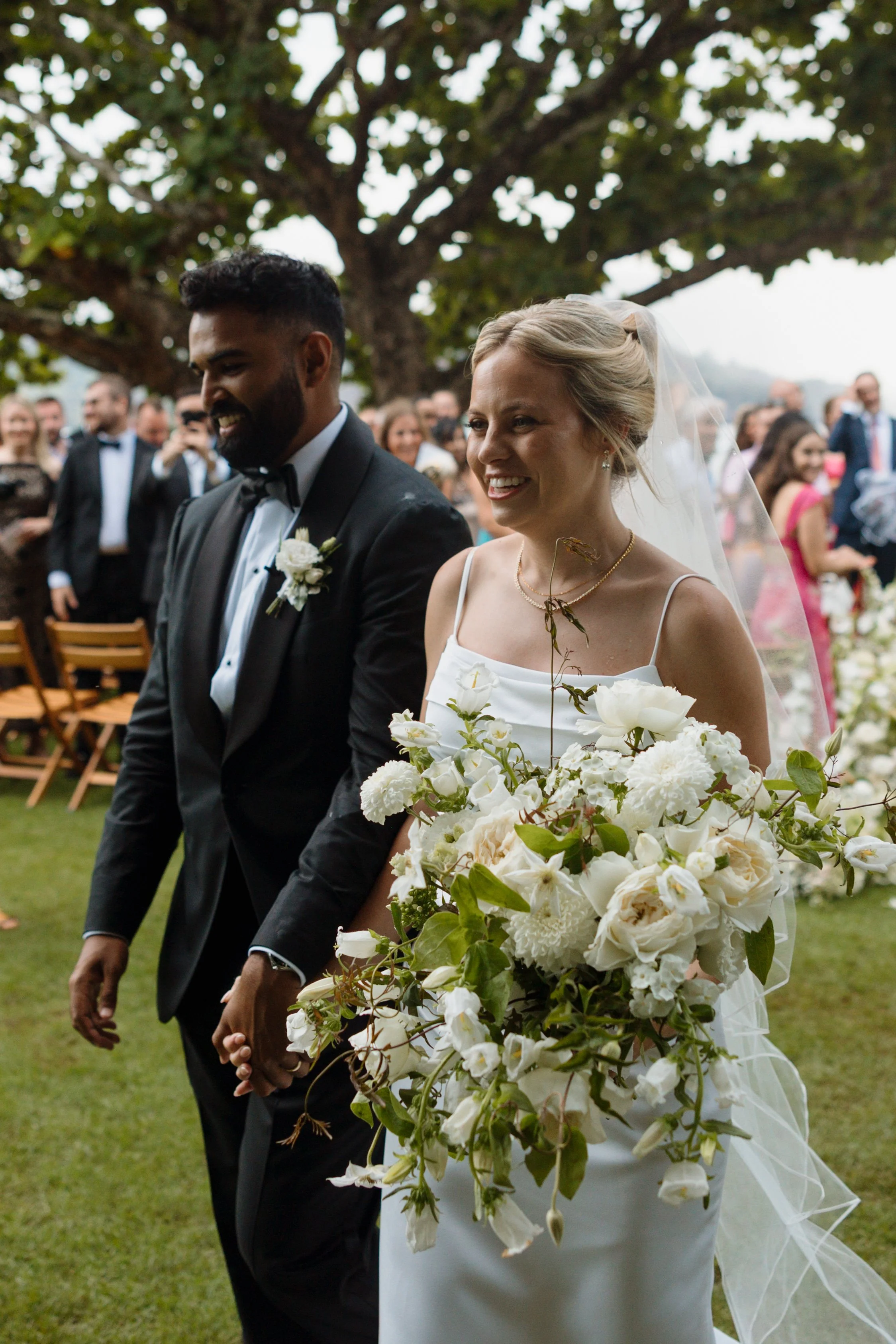 Bride and groom holding hands during wedding ceremony outdoors, with guests in background, under a large tree.