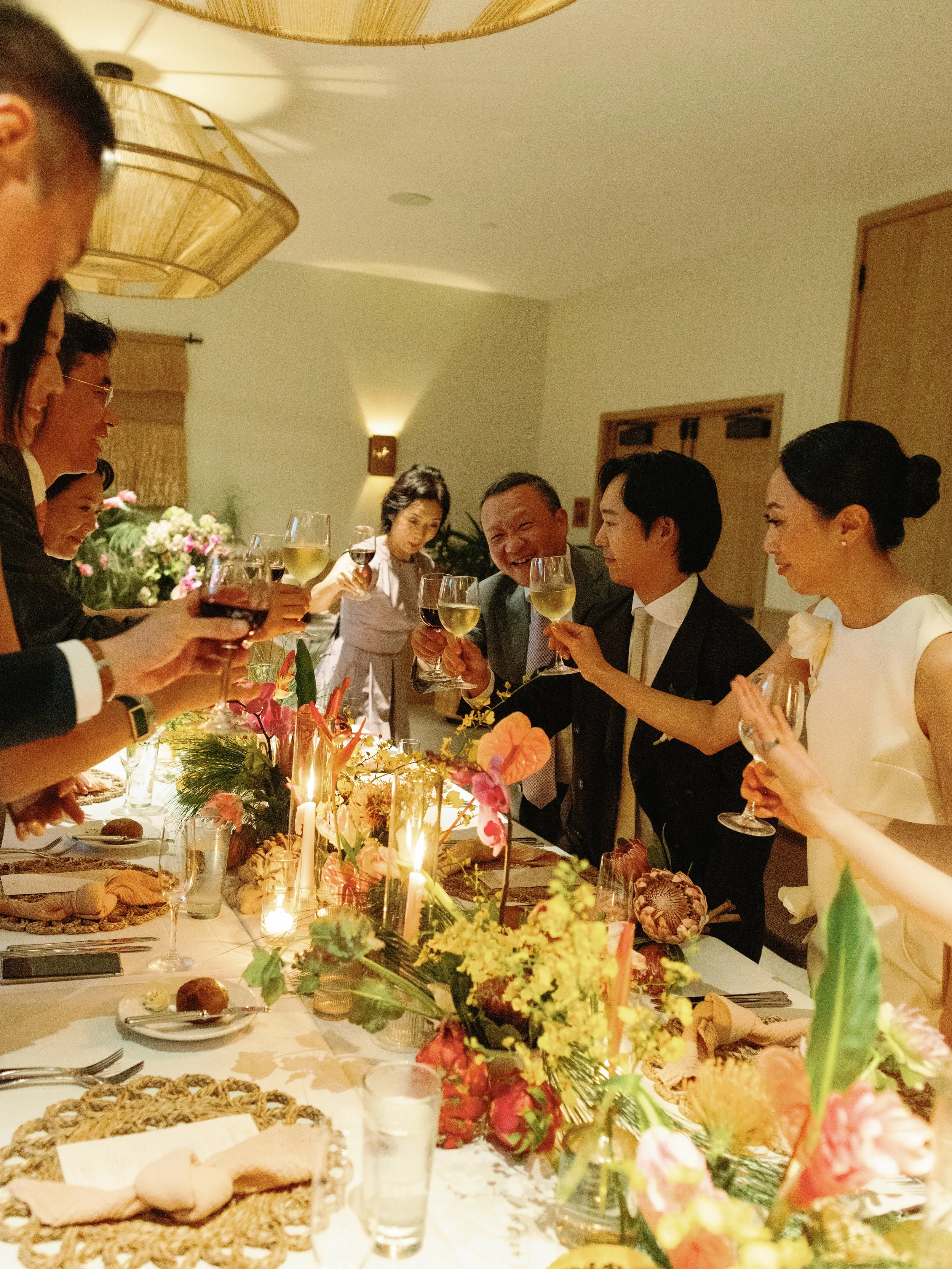 People raising glasses in a toast at a dinner party with a decorated table featuring flowers and candles.