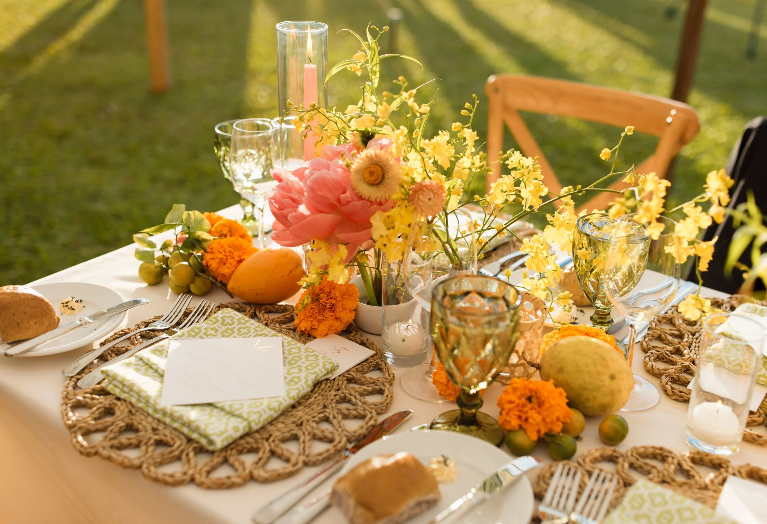A beautifully set outdoor dining table decorated with pink, yellow, and orange flowers, candles, and elegant glassware, with a grassy background and wooden chairs.