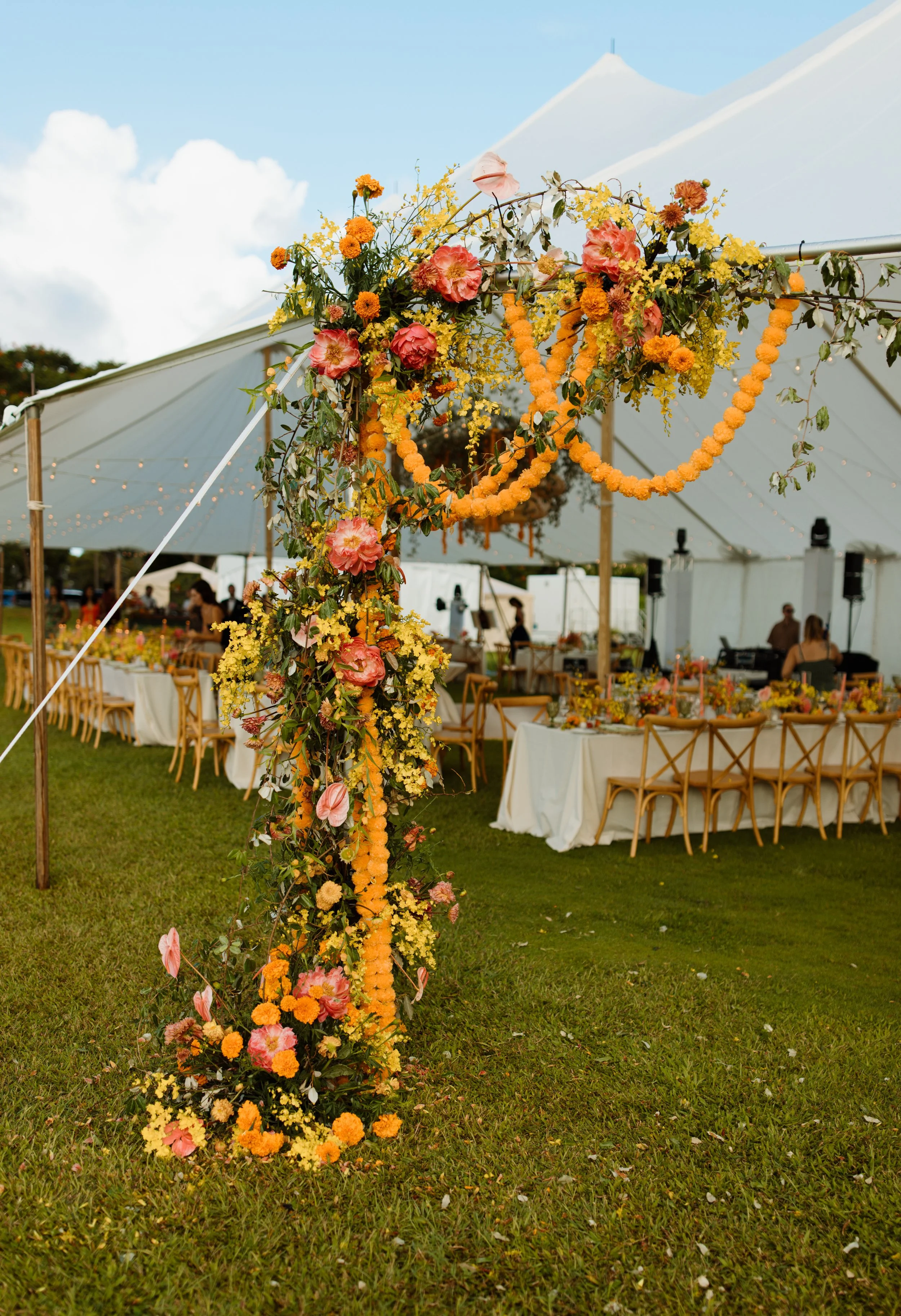 Decorated wedding arch with yellow, orange, and pink flowers, set against a white tent with tables and chairs underneath, on a grassy outdoor area.