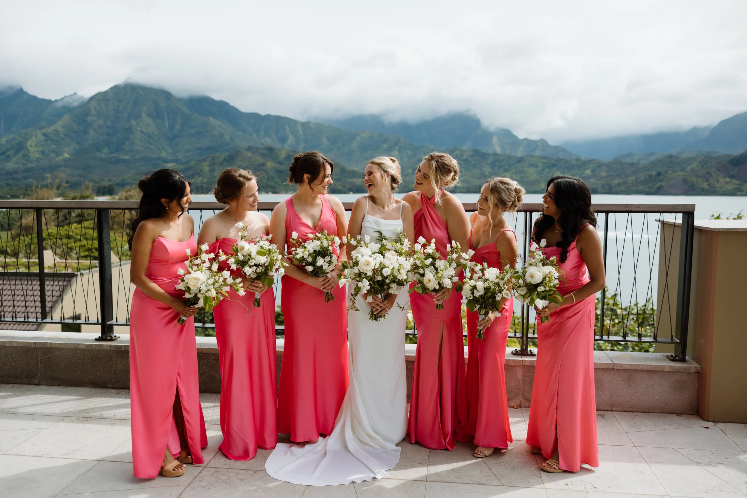 A group of seven women dressed in pink and white dresses holding bouquets of white flowers, standing on a balcony with a mountain landscape in the background.