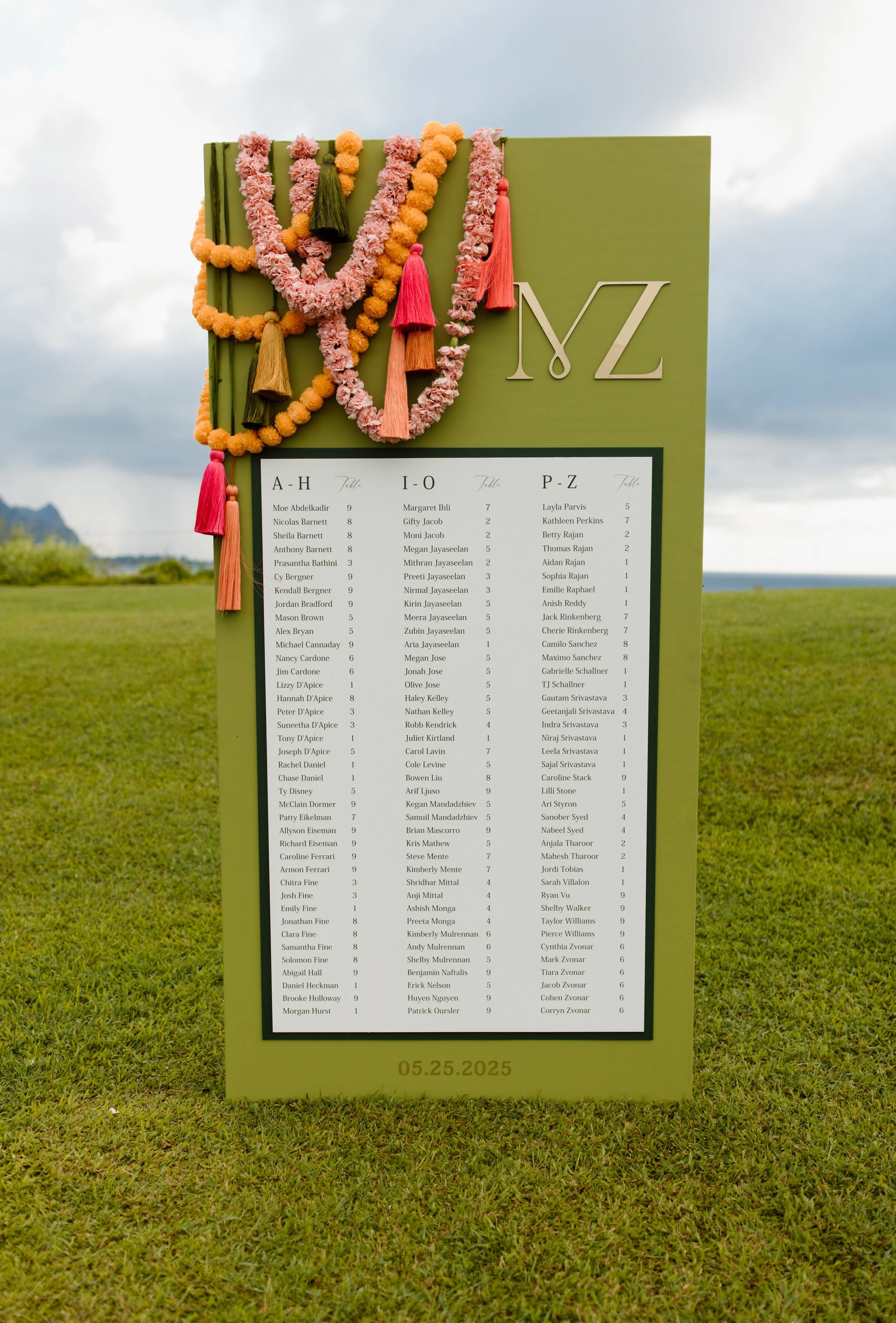 Graduation ceremony program board decorated with pink, orange, red, and green floral garlands and tassels, displaying a list of names, tables, and a date of May 25, 2025, outdoors.