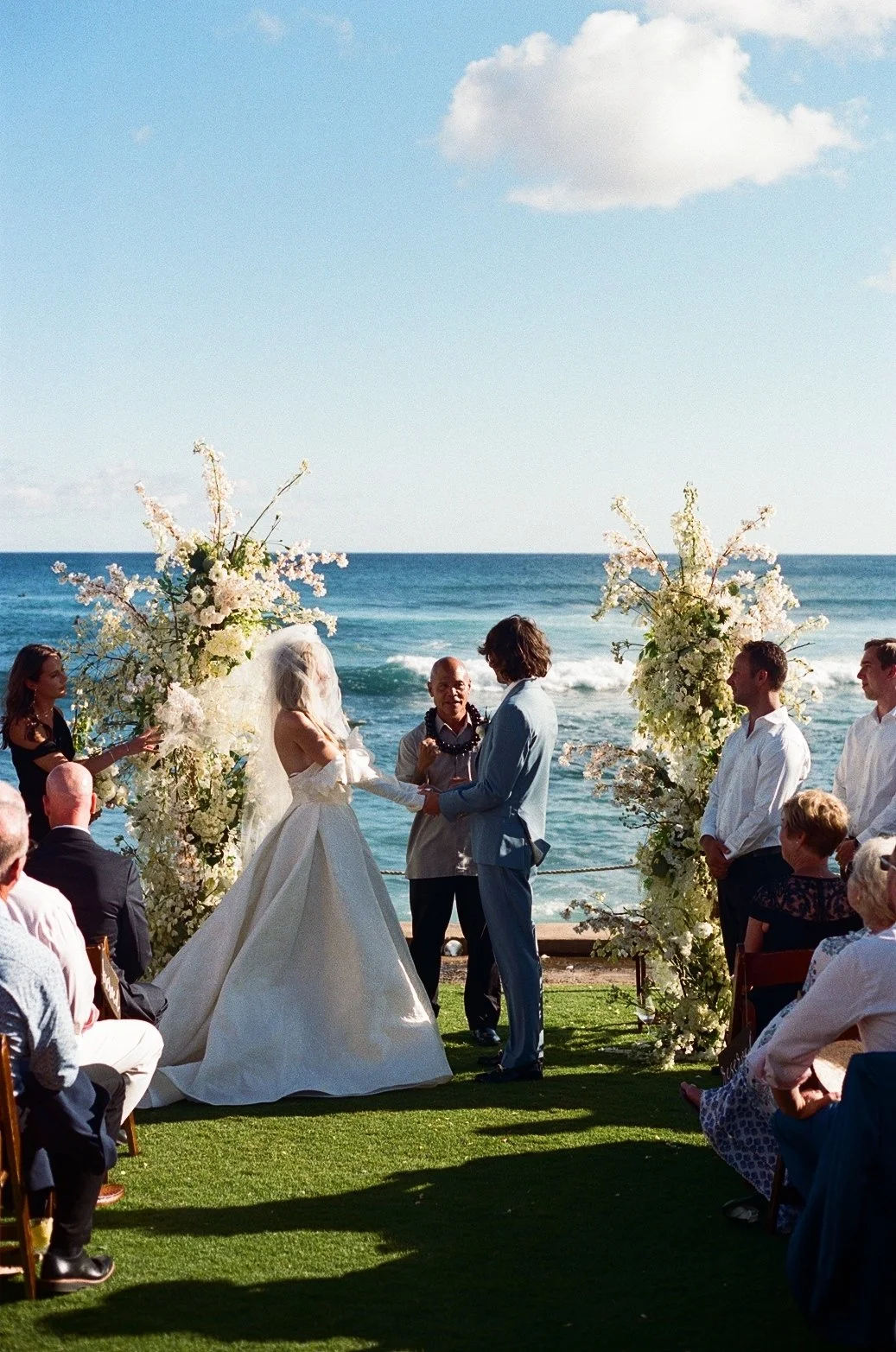 A wedding ceremony taking place outdoors by the ocean, with the bride and groom holding hands and exchanging vows, surrounded by floral decorations and seated guests.