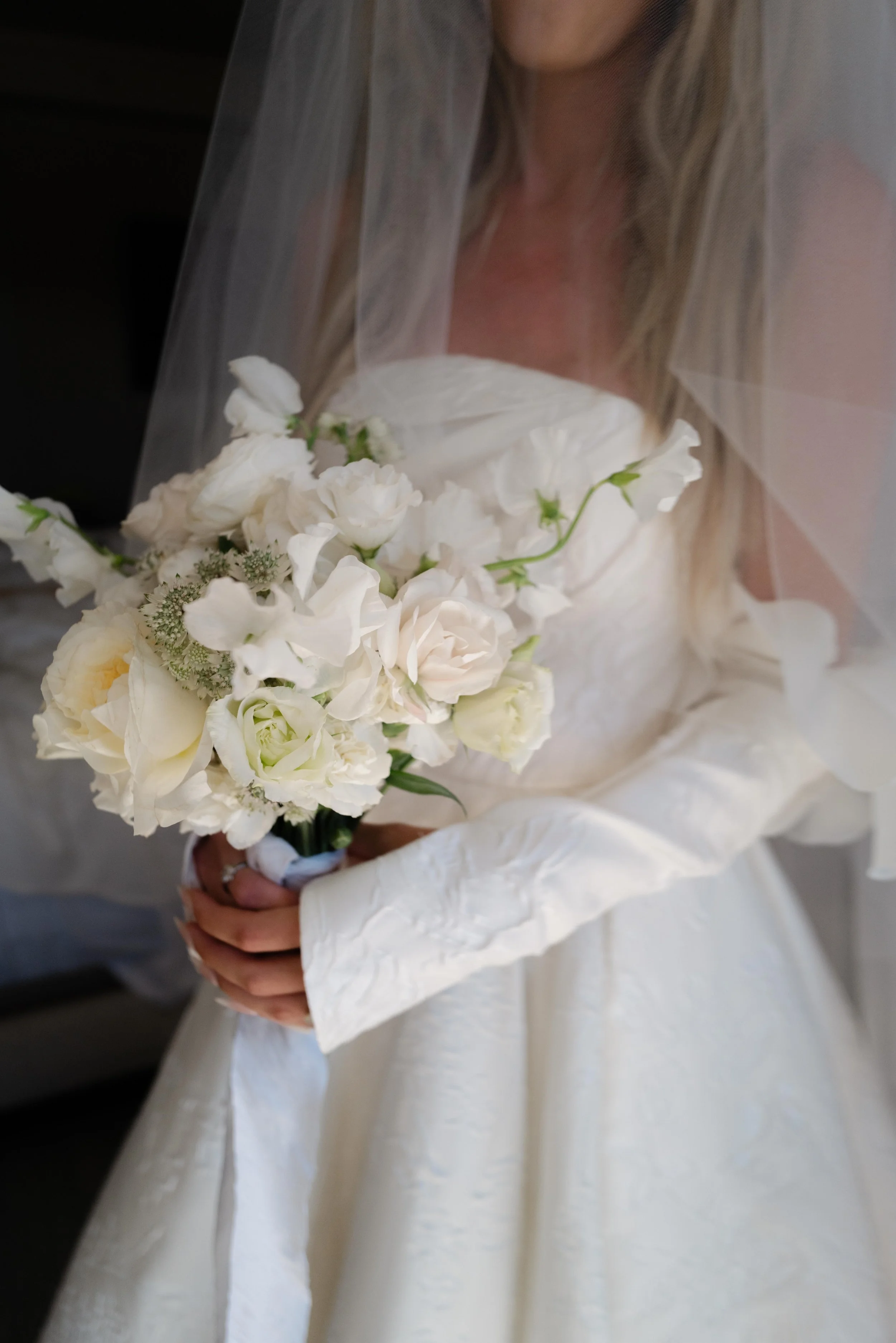 A woman in a white wedding dress holding a bouquet of white flowers with greenery, standing behind a white veil.