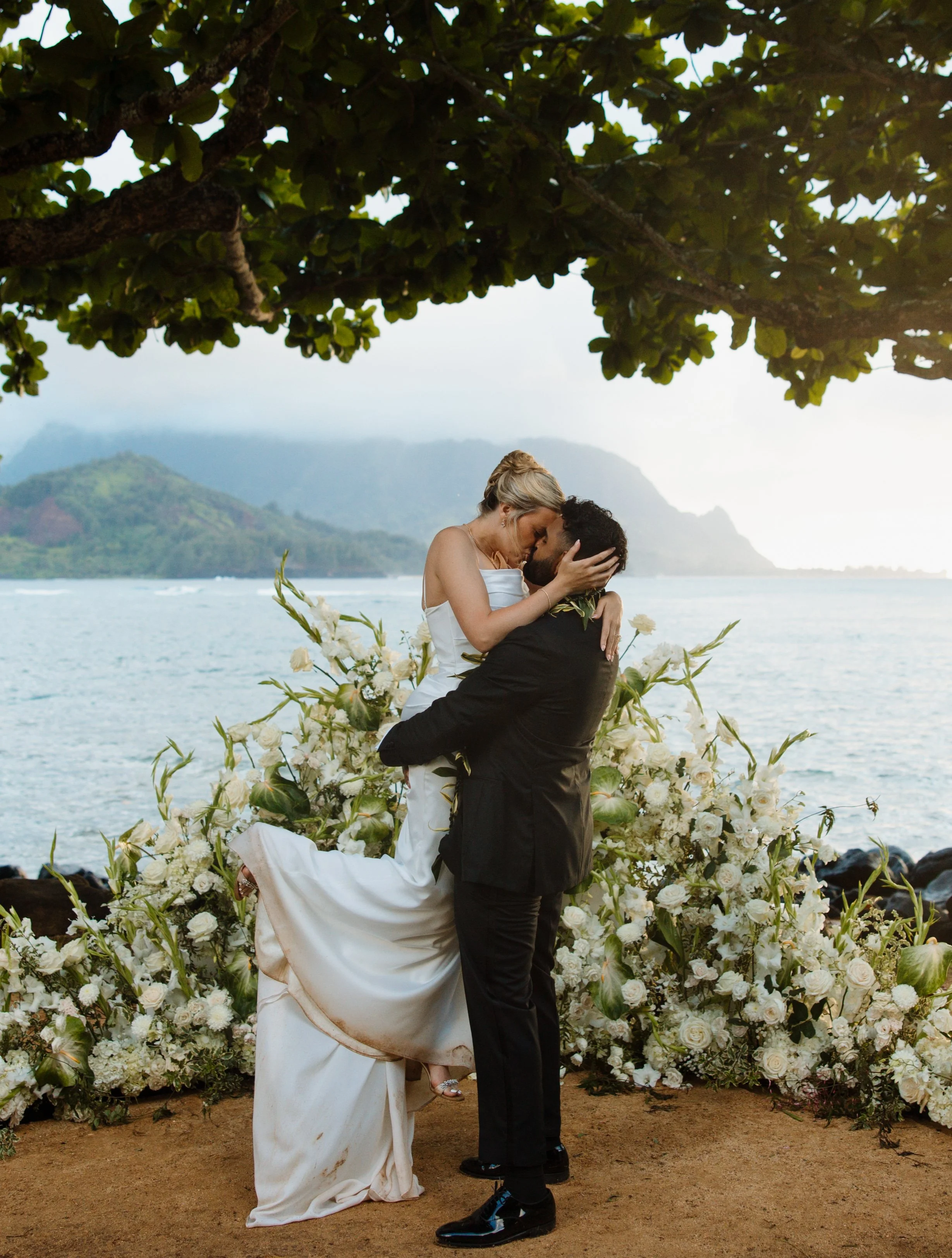 A couple is kissing on a beach during their wedding ceremony, with the bride wearing a white dress and the groom in a black suit, surrounded by white flowers and overlooking the ocean with mountains in the background.