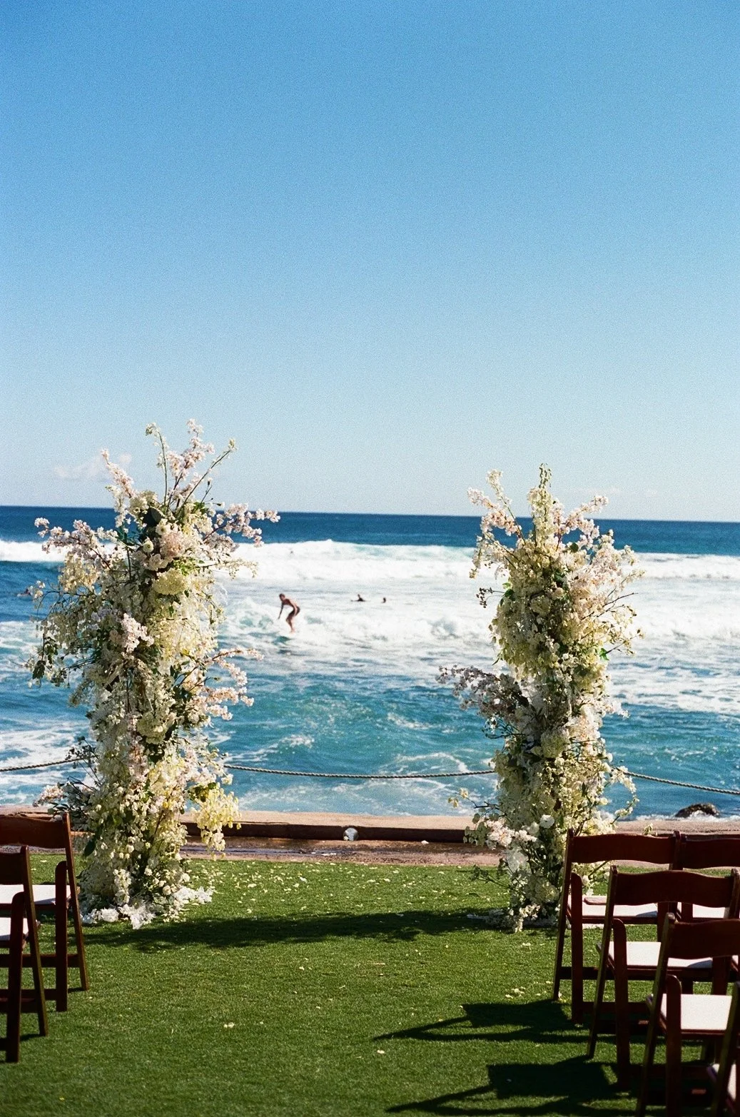 A seaside wedding setup with a floral arch made of white and pale pink flowers and greenery on a grassy area, overlooking the ocean and surfers in the water, with blue sky overhead.