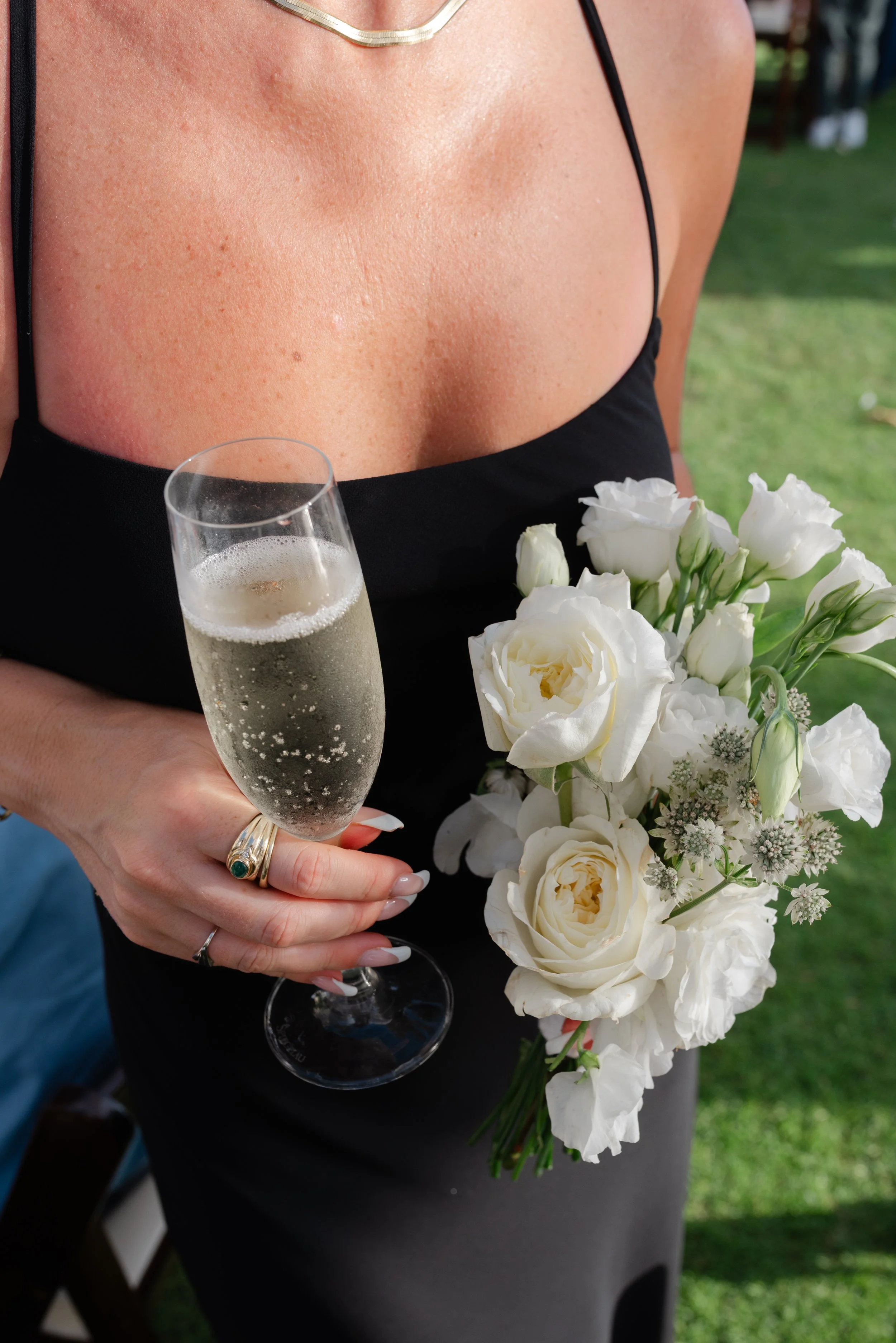 A woman holding a bouquet of white flowers and a glass of champagne, wearing a black dress and a gold necklace, outdoors in a garden setting.