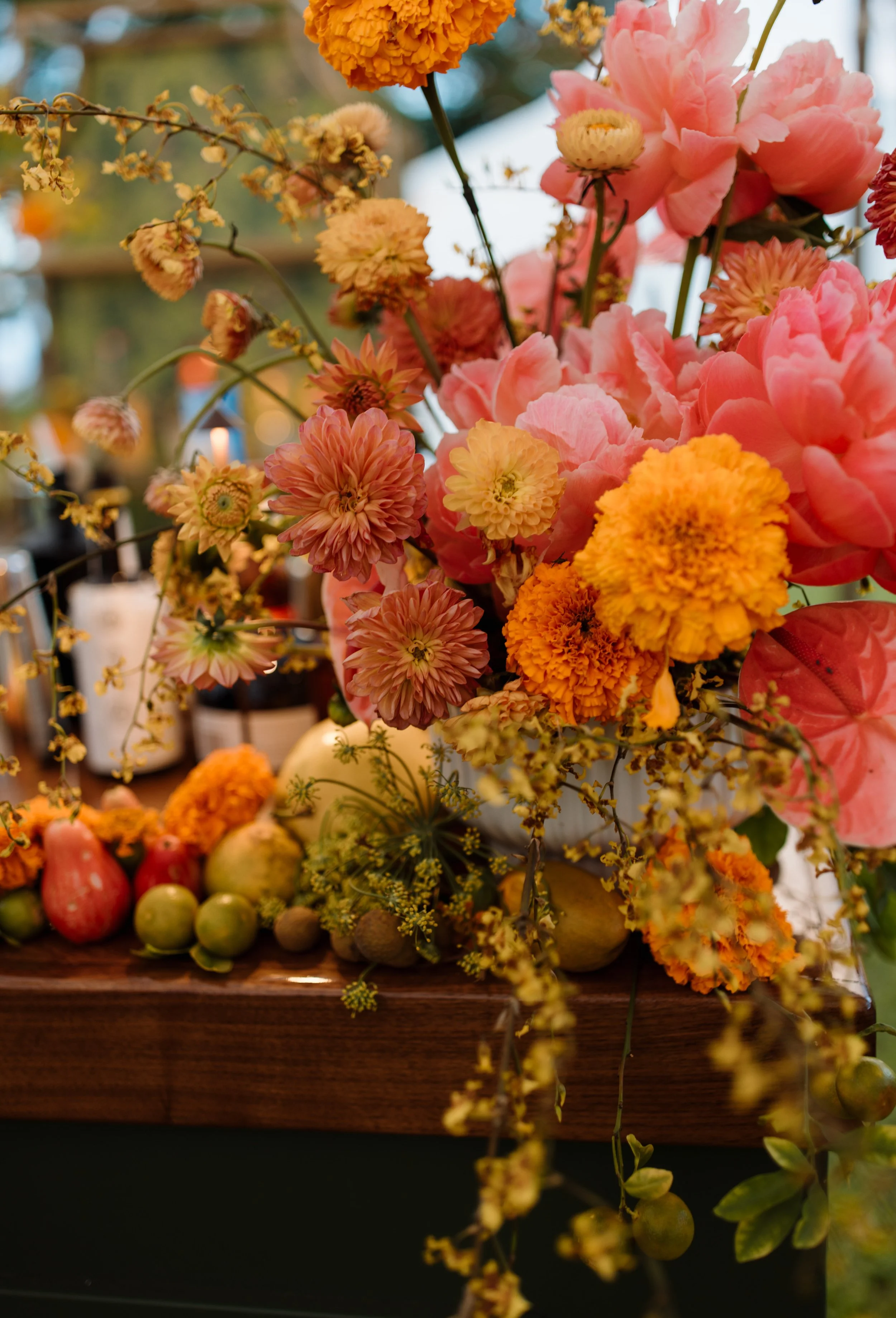 Close-up of a floral arrangement with pink, orange, and yellow flowers, and fruit such as pears and figs on a wooden surface.