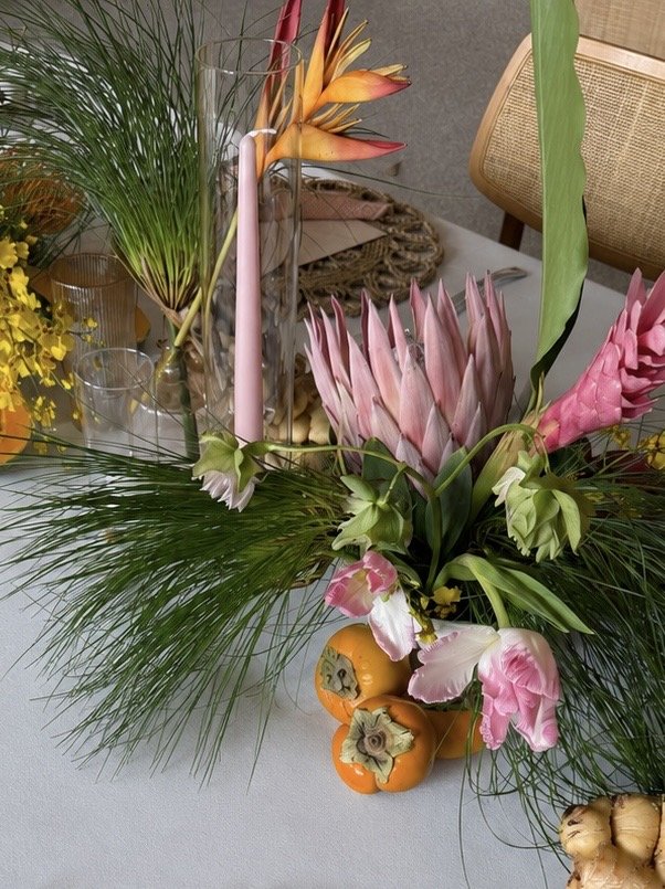 A floral arrangement on a table featuring pink and white flowers, green leaves, and decorative peppers, with a glass candle holder containing a pink taper candle.