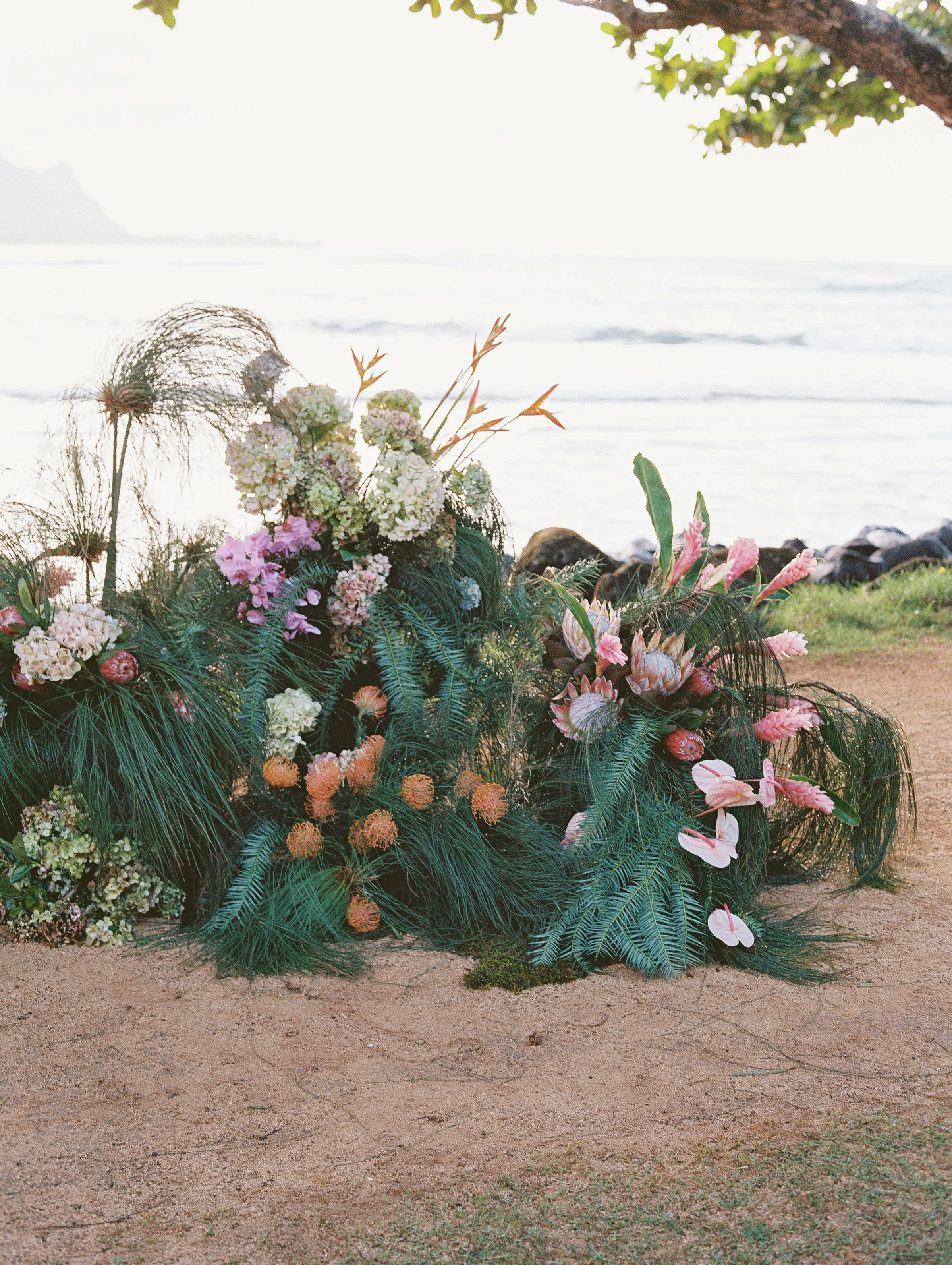 Colorful floral arrangement with pink, white, and orange flowers and lush green foliage, placed on a sandy outdoor surface near a body of water, with a tree branch and leaves overhead.