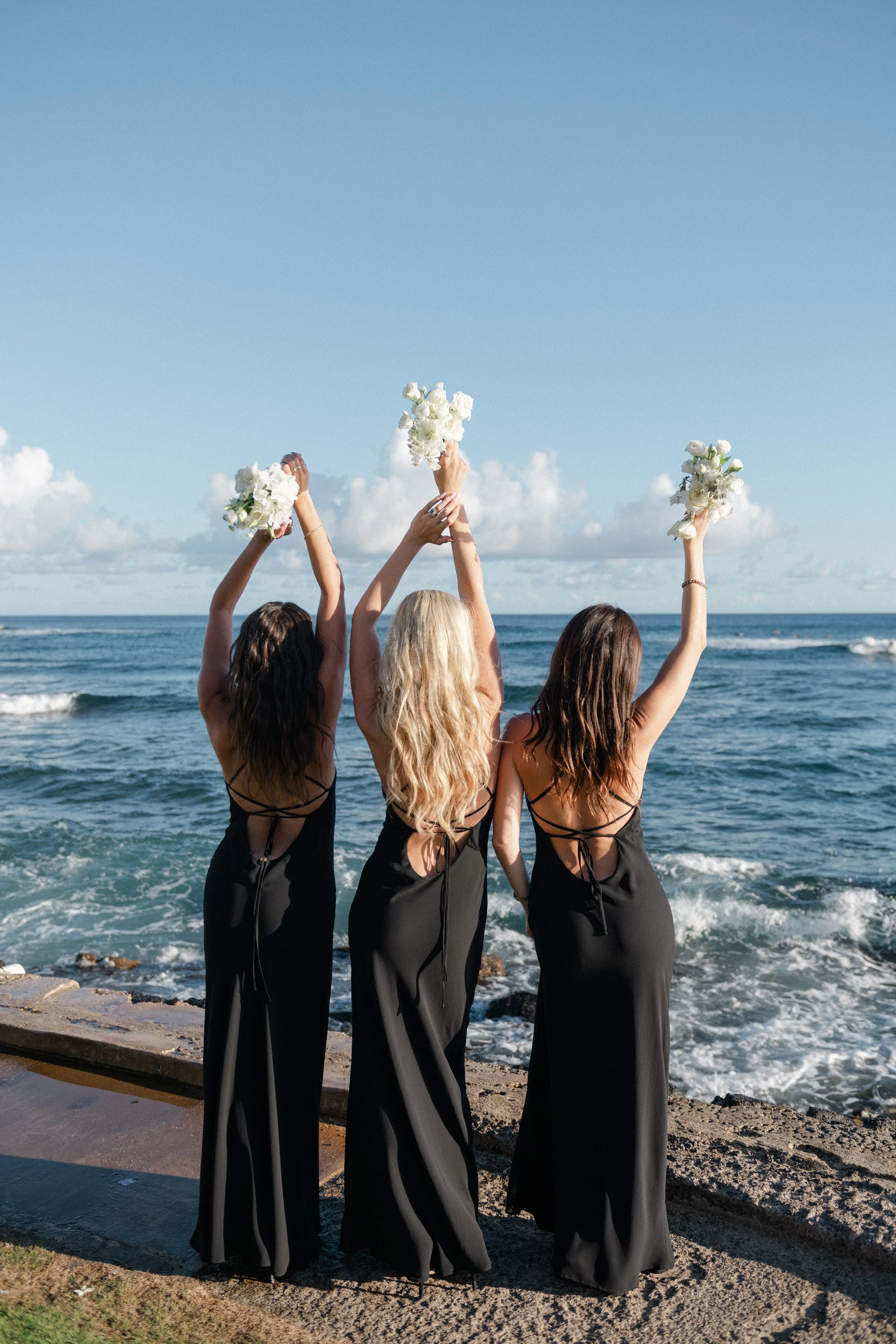 Three women in black dresses standing on a beach with their backs to the camera, holding white flower bouquets high in the air, facing the ocean during daylight.