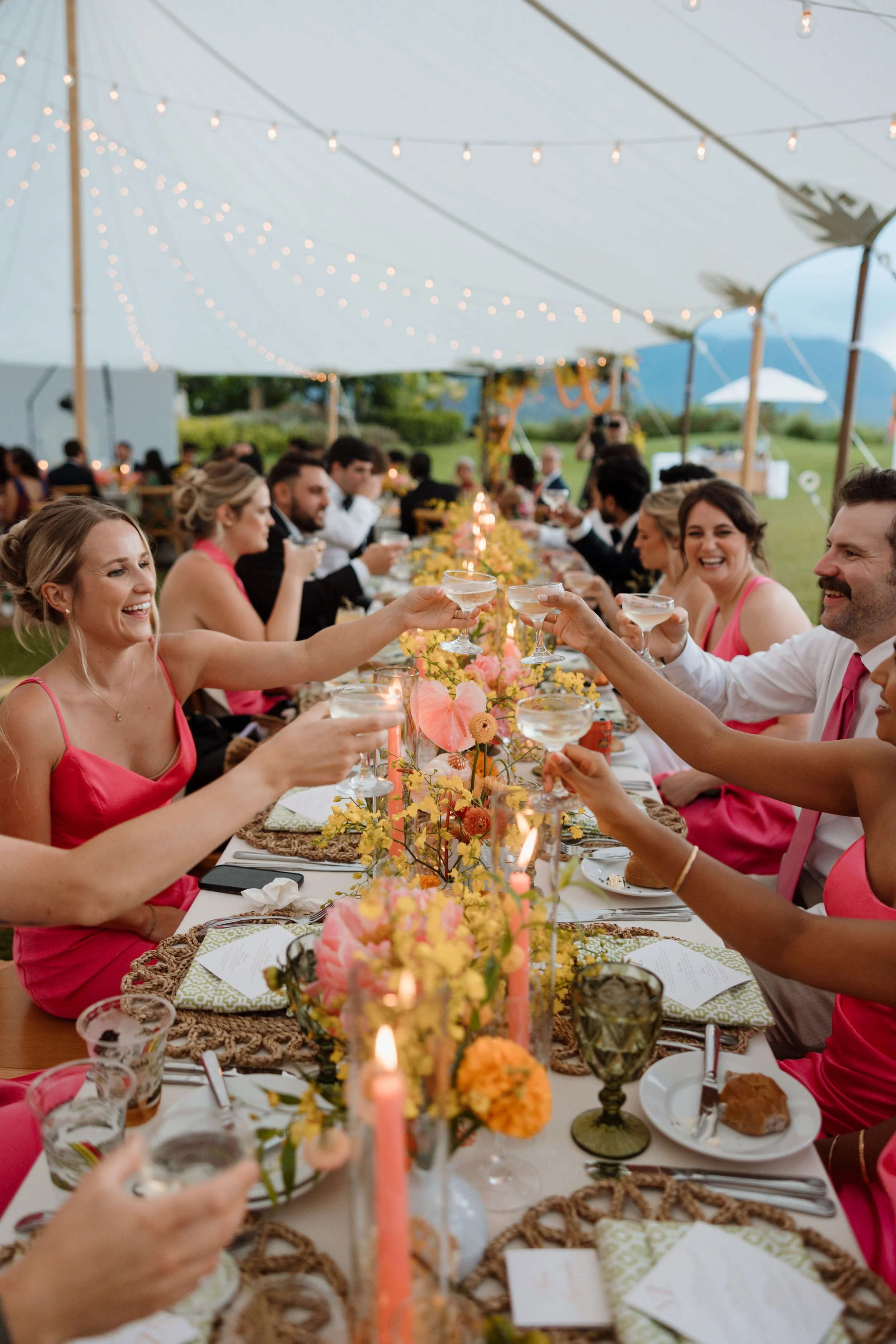 People sitting around a long dinner table under a white canopy at an outdoor gathering during sunset, enjoying drinks and conversation, decorated with flowers and string lights.