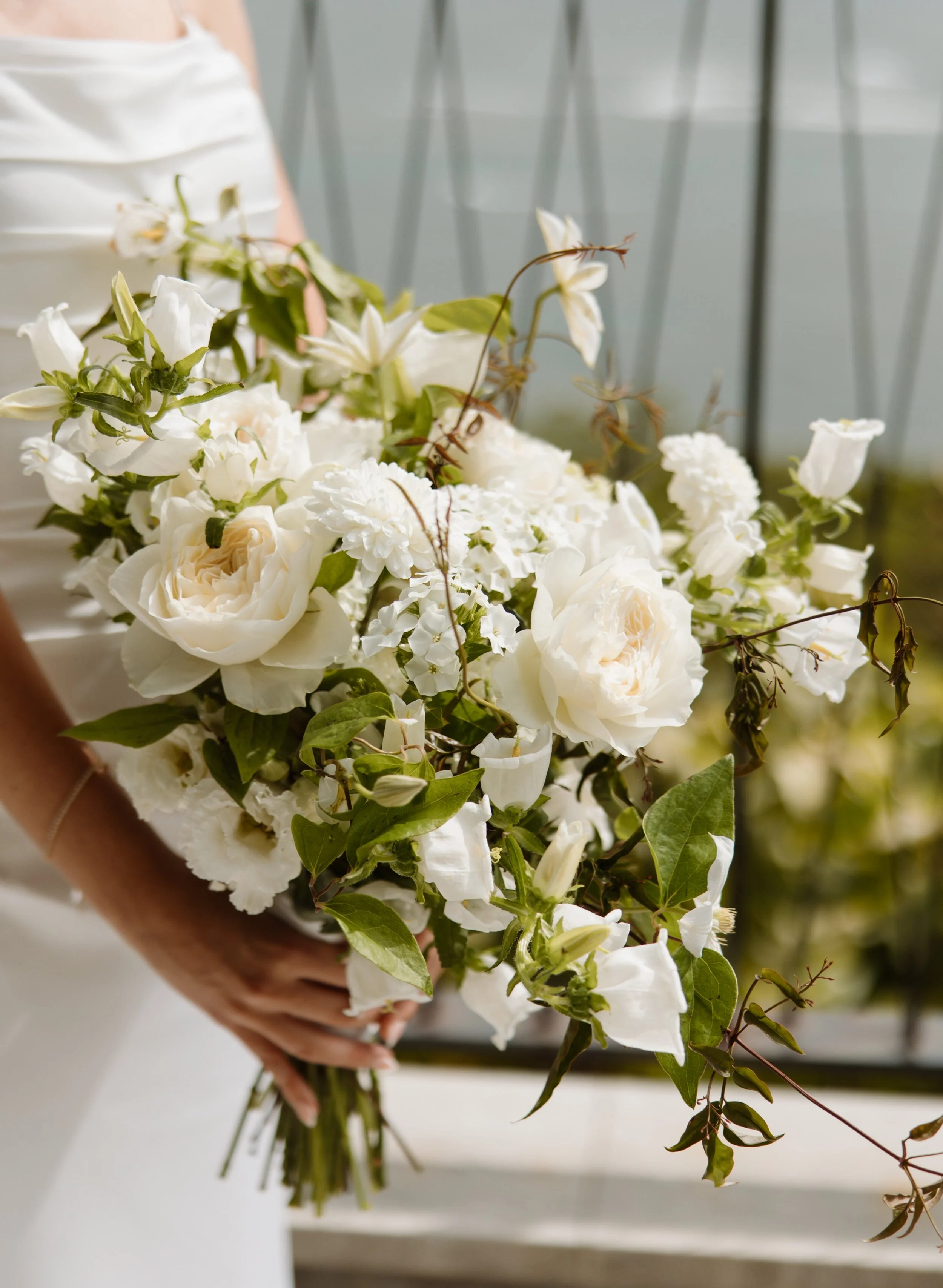 A person in a white dress holding a bouquet of white flowers with green leaves, standing in front of a light-colored wall with a gray railing.