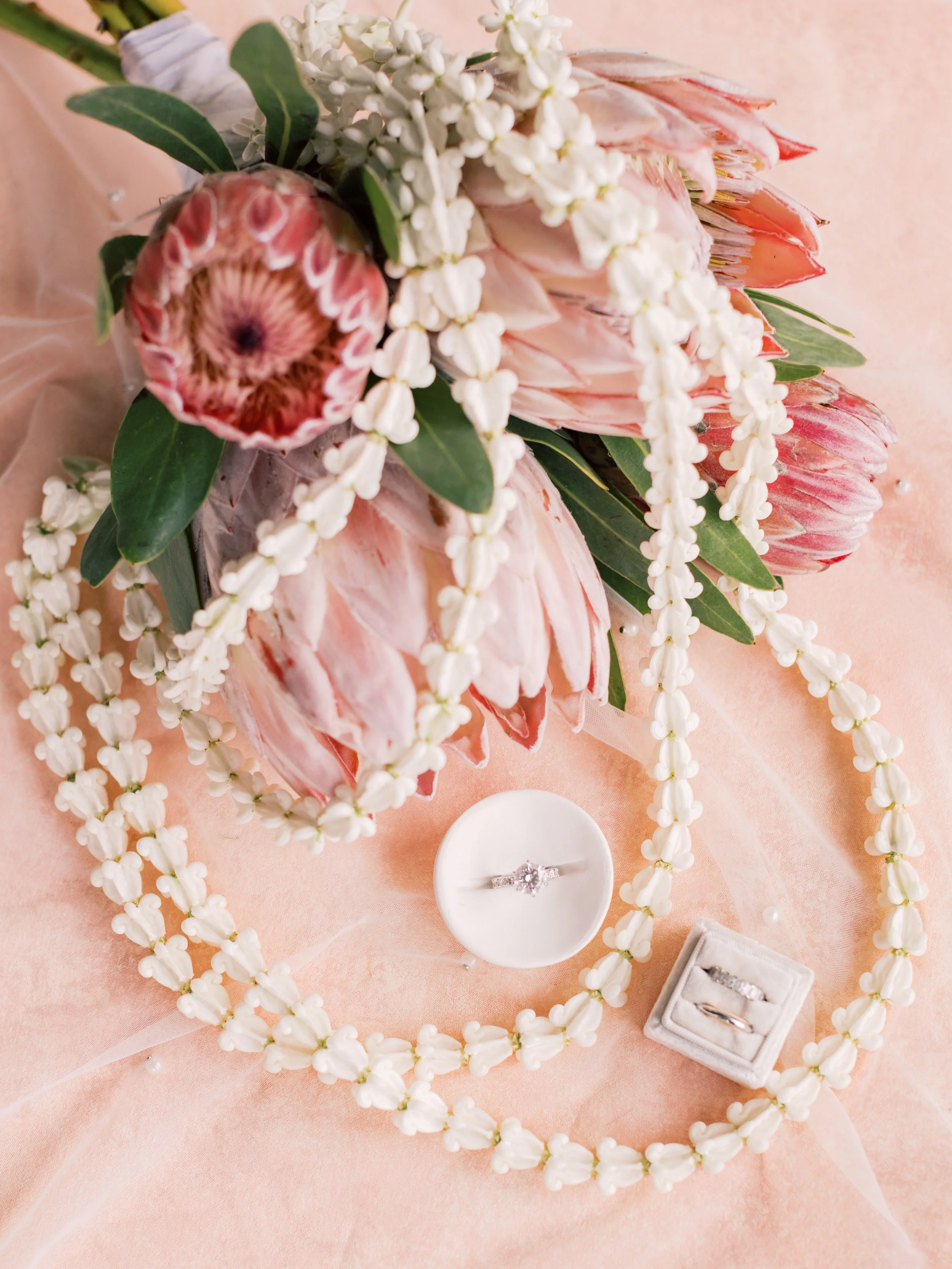 A floral arrangement with pink protea flowers, white accent flowers, green leaves, and white beads arranged on pink fabric. A white ring box with a diamond engagement ring and a matching wedding band is also visible.