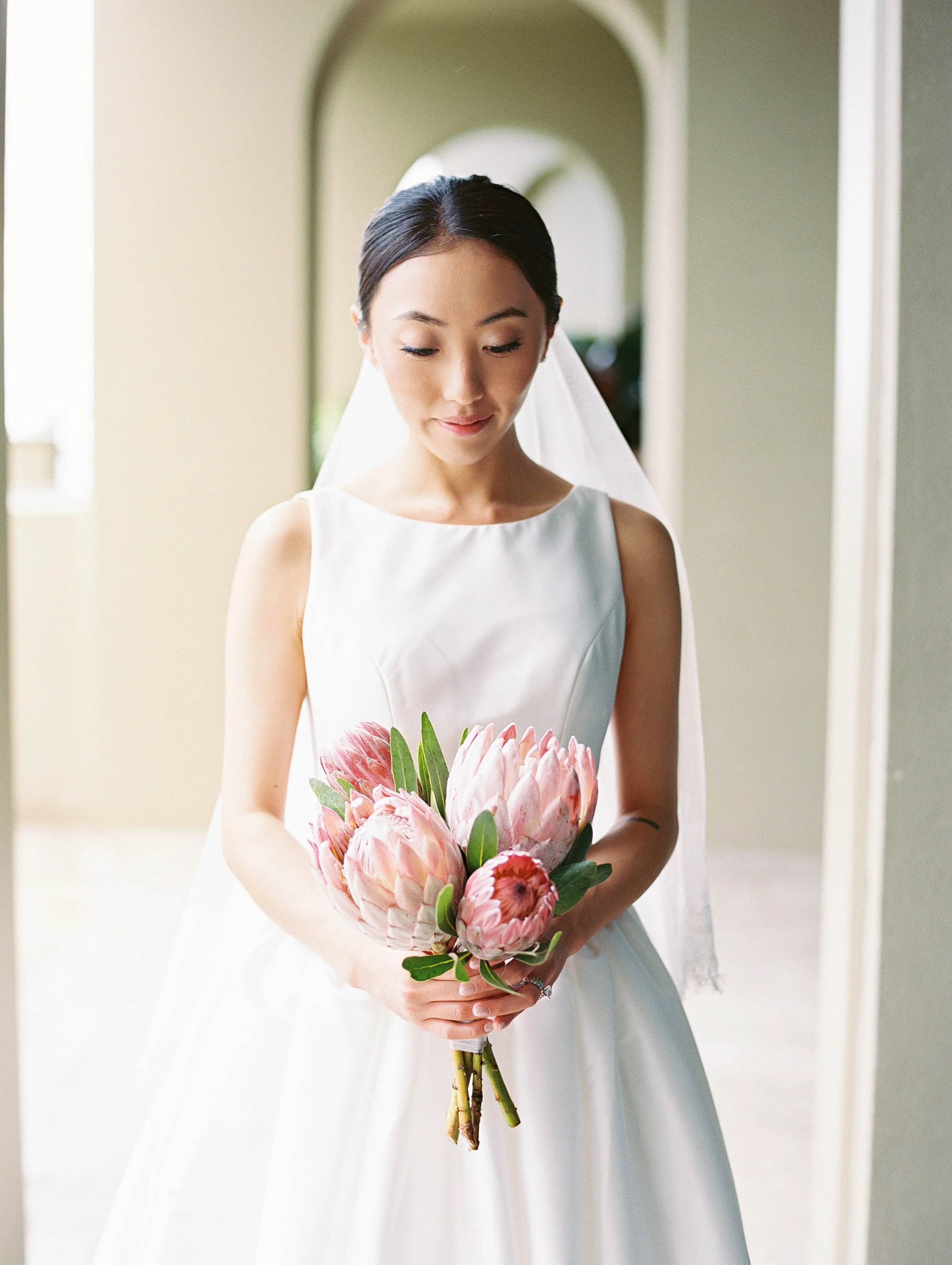 A bride in a white wedding dress holding a bouquet of pink and white flowers, standing in front of an arched doorway.