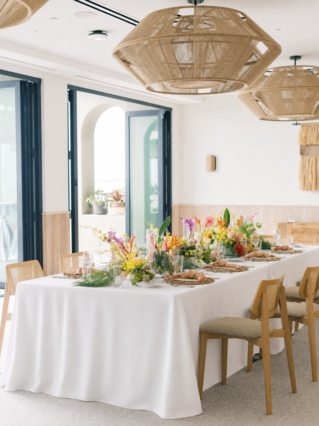 A long dining table decorated with a white tablecloth and a colorful flower arrangement, set with beige napkins, glassware, and plates in a bright, modern room with large windows and wooden accents.