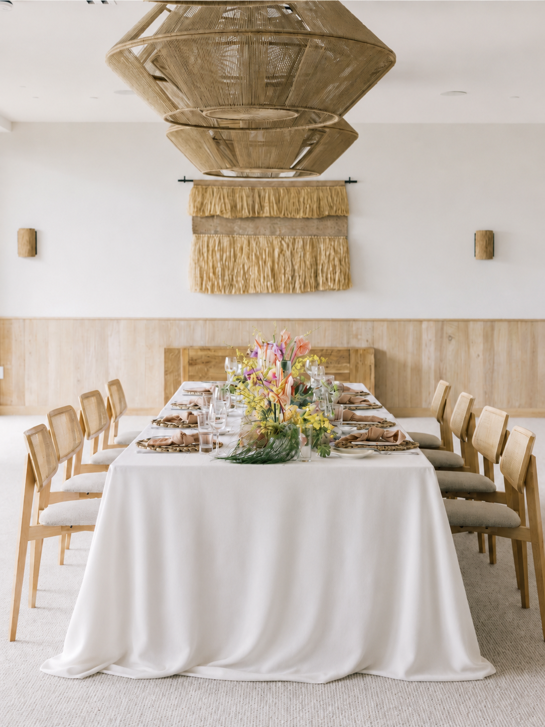 A dining room set for a formal event with a long white tablecloth, floral centerpiece, and place settings. Wooden chairs surround the table, and a decorative wall hanging and a large woven chandelier are visible on the wall.