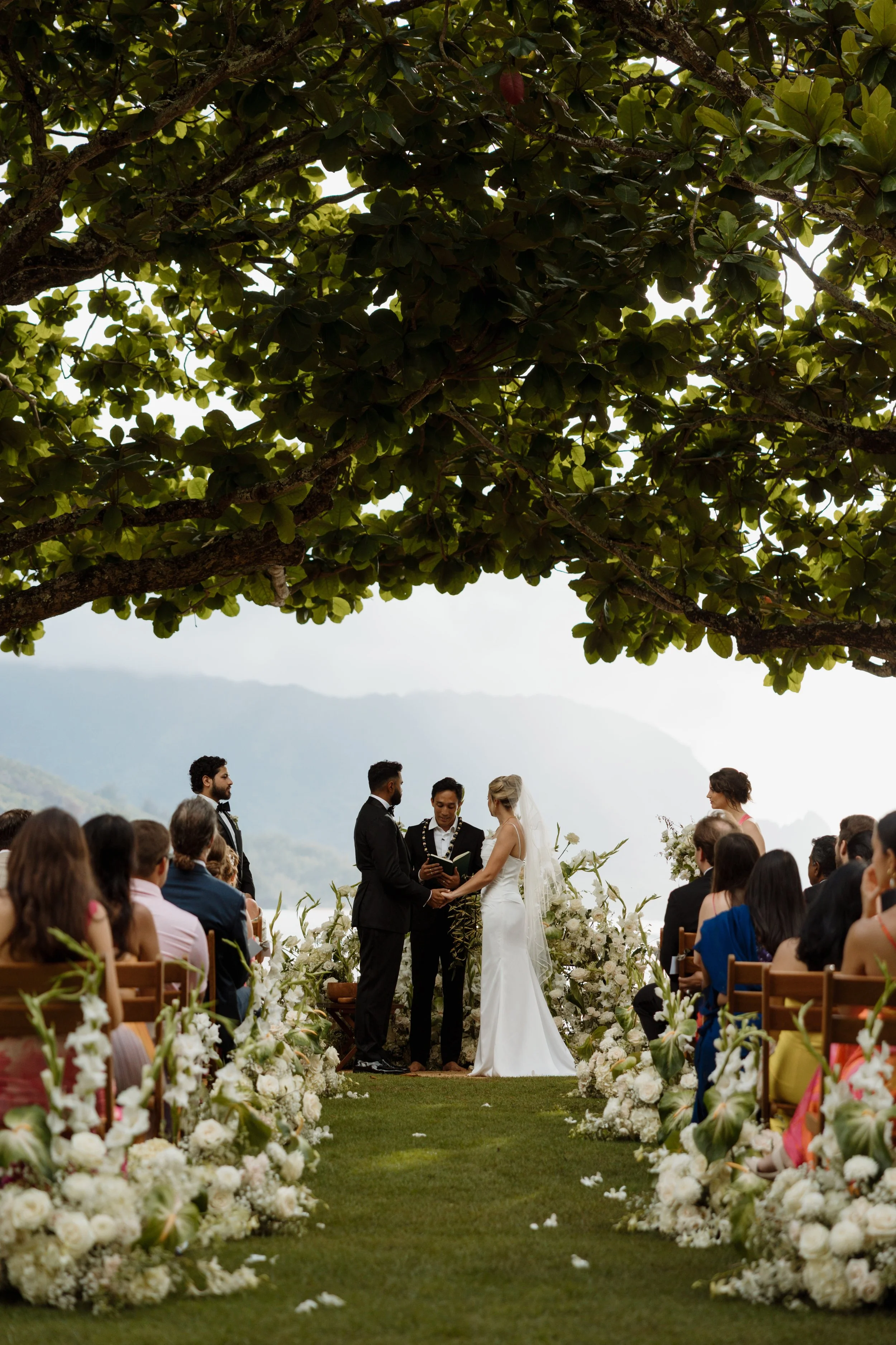 A wedding ceremony happening outdoors with an altar decorated with white flowers, under a large leafy tree. The bride and groom are holding hands and exchanging vows, surrounded by seated guests. A mountainous landscape is visible in the background.