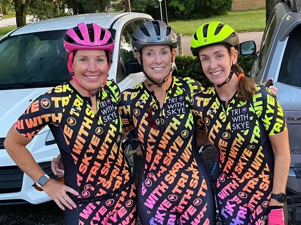 3 women posing together in bike helmets and race suits that read "Tri with Skye"