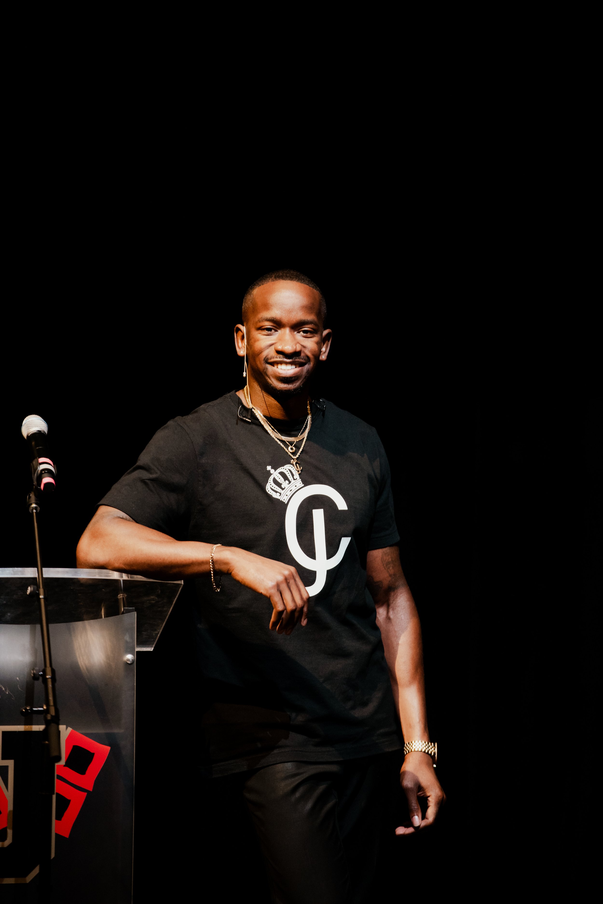 “Charleston Girley smiling beside a podium with a microphone, wearing a black logo shirt against a dark background.”