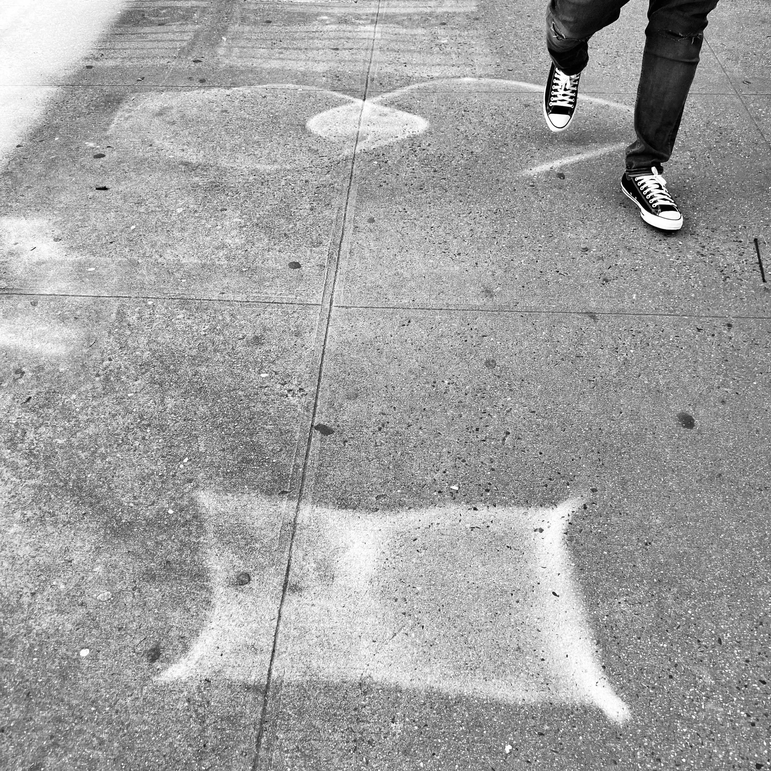 A black and white photo of a sidewalk with spray-painted white stars and circles, with part of a person wearing jeans and black and white sneakers walking through the scene.