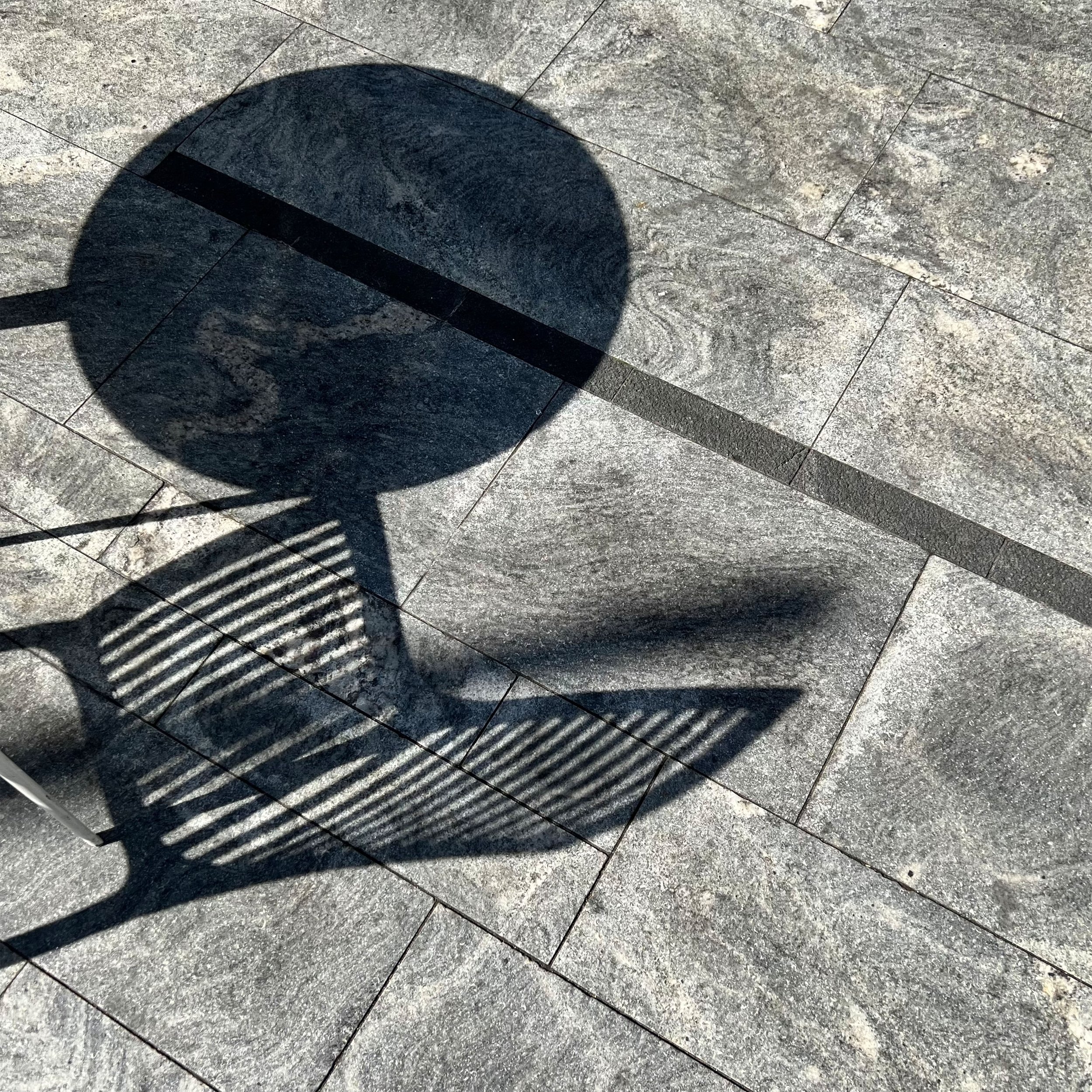 Shadow of a chair with a circular backrest cast on a tiled stone floor, with sunlight creating striped shadow on the chair.