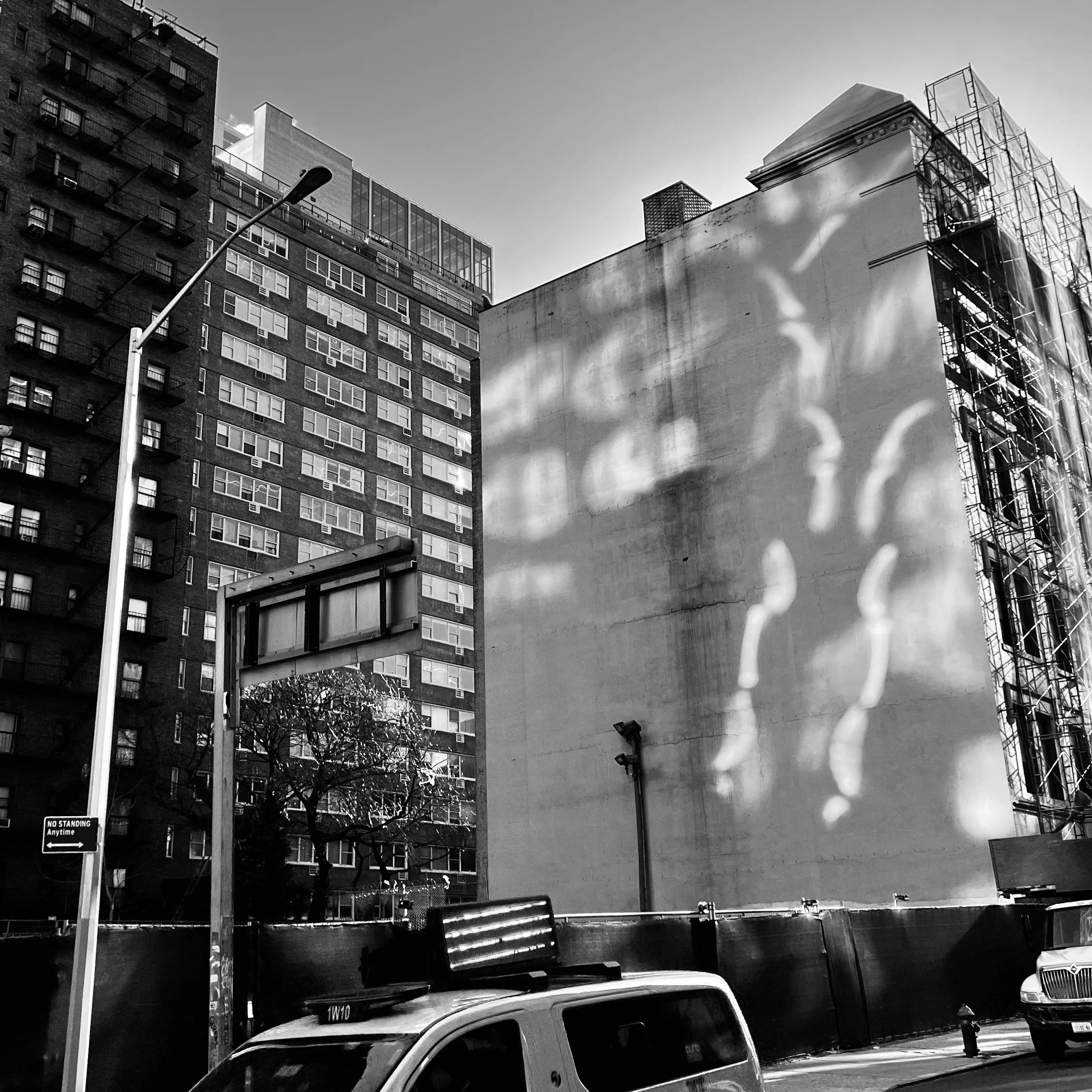 Urban cityscape in black and white with tall buildings, a streetlamp, a tree, vehicles including a taxi, and a building with scaffolding. Shadows of hands are projected onto a blank wall.