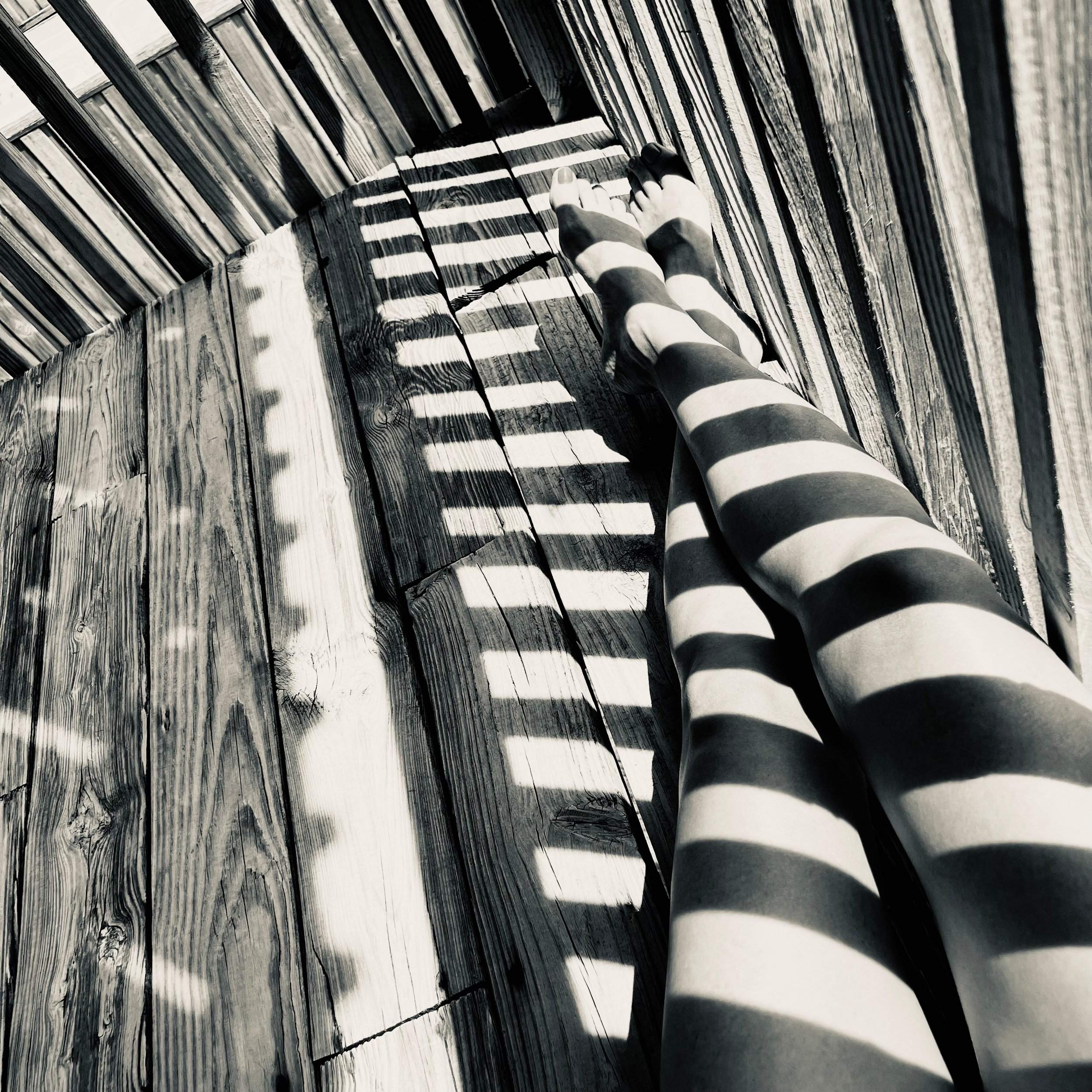 A person's legs and feet wearing striped tights, sitting on a wooden deck, with shadows from a slatted fence creating striped patterns on the legs and the deck.