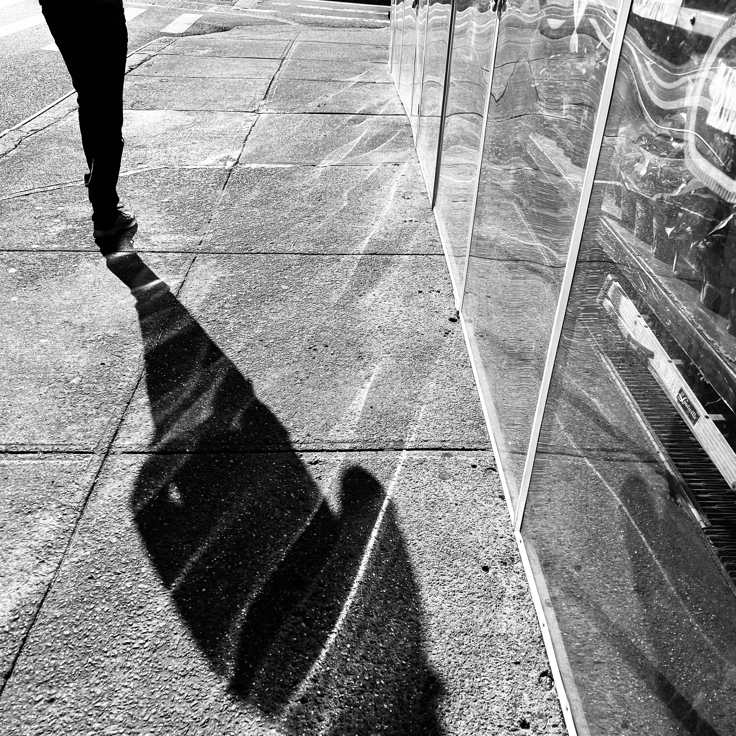 A sidewalk scene in black and white showing a person walking away, carrying a suitcase. There are shadows cast on the sidewalk, and a storefront with reflective glass windows on the right.