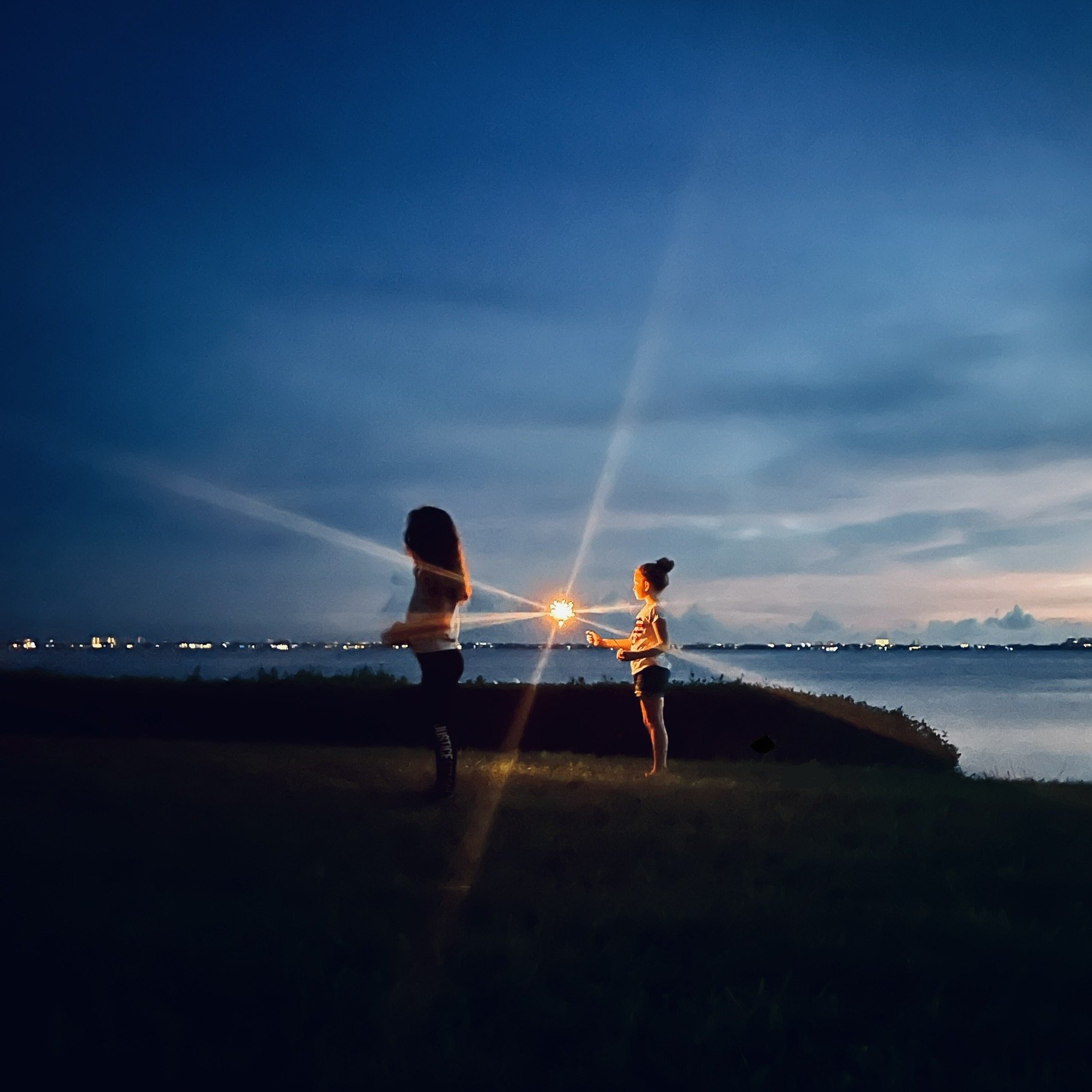 Two women standing outdoors near water at sunset, one using a phone, with the setting sun positioned between them, creating a starburst effect.
