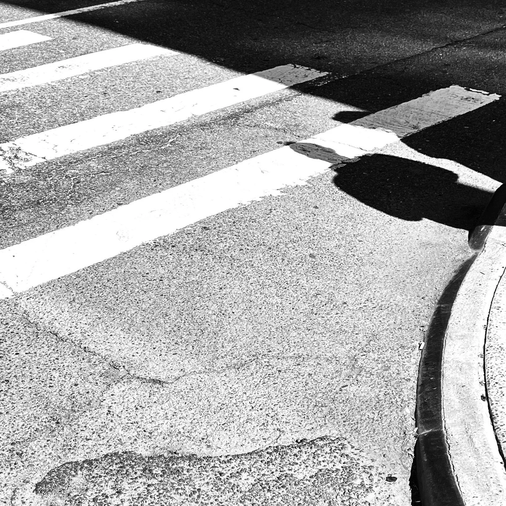 Black and white photo of a crosswalk with white striped lines on the asphalt. Part of a curb with a rounded corner and shadow of a street sign are visible while sunlight creates sharp shadows.