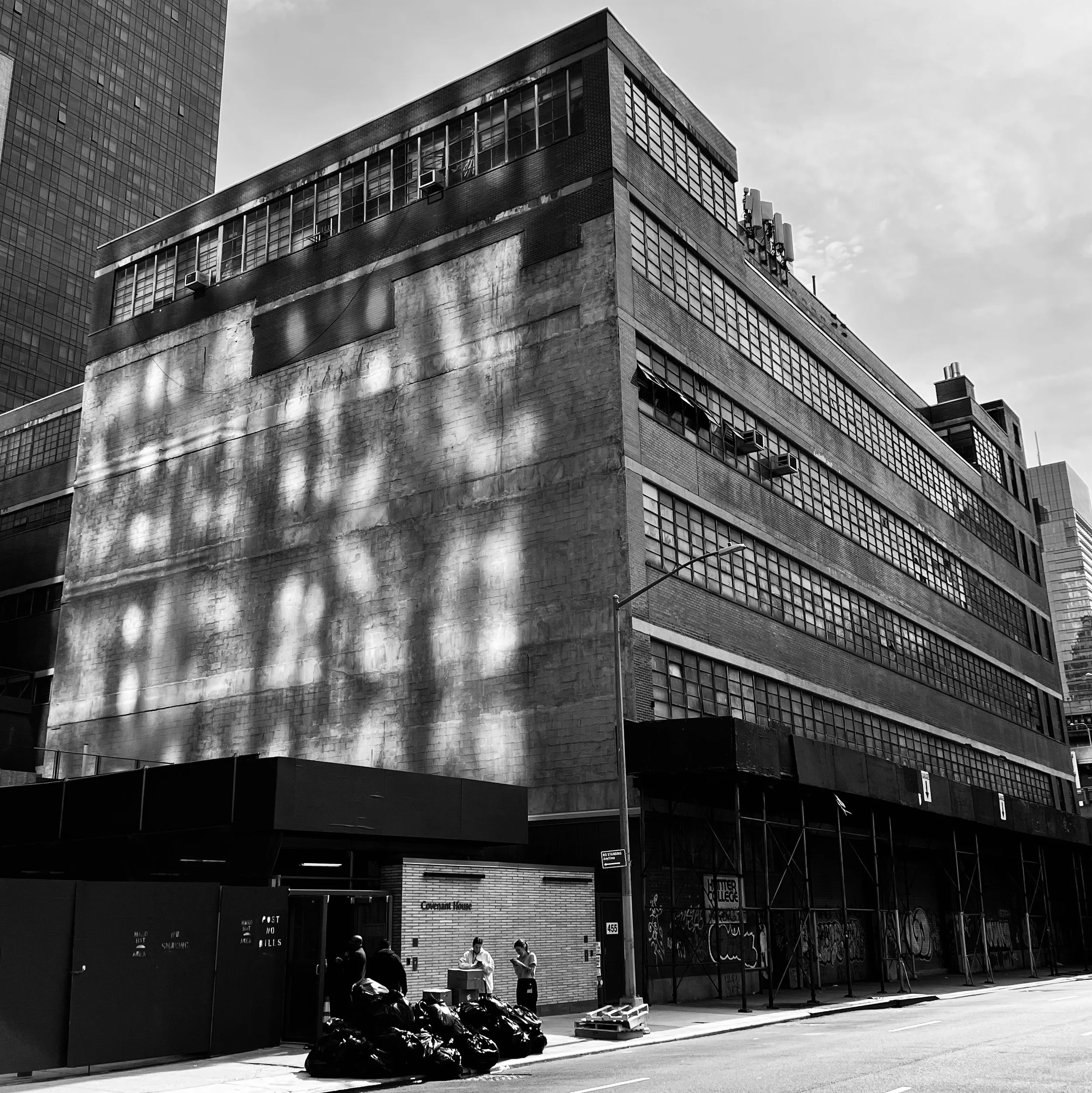 Black and white photo of a multi-story urban building with a large blank wall on one side, shadow patterns on the wall, and scaffolding at the base. People are standing near the entrance, and garbage bags are piled nearby.